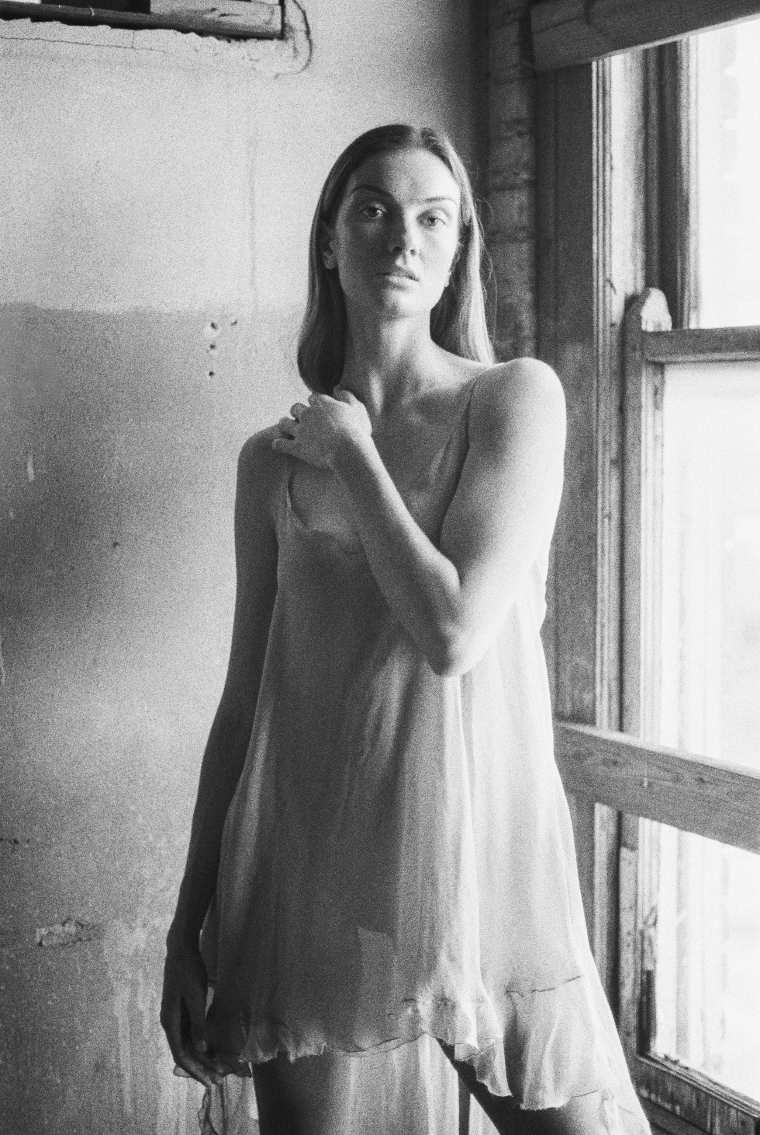 Black and white photo of a woman with long hair, wearing a dress, standing in front of a window in a rustic room, looking at the camera.