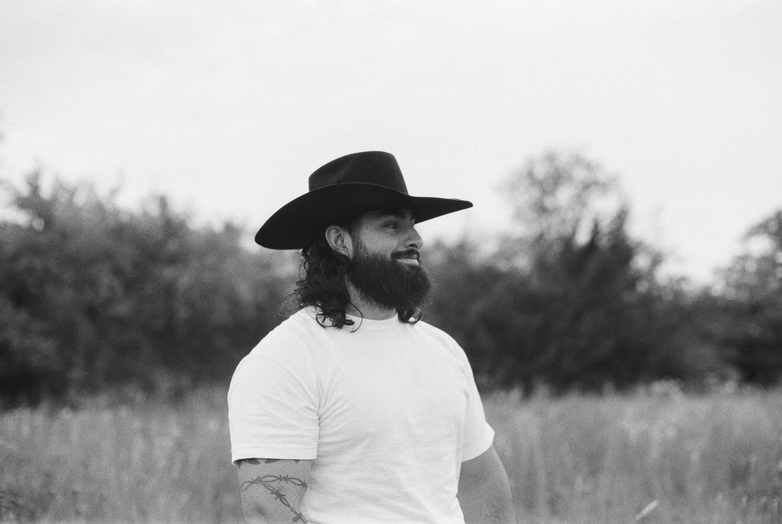 A man with a beard and long hair wearing a wide-brimmed hat and a white T-shirt outdoors with trees and grass in the background.