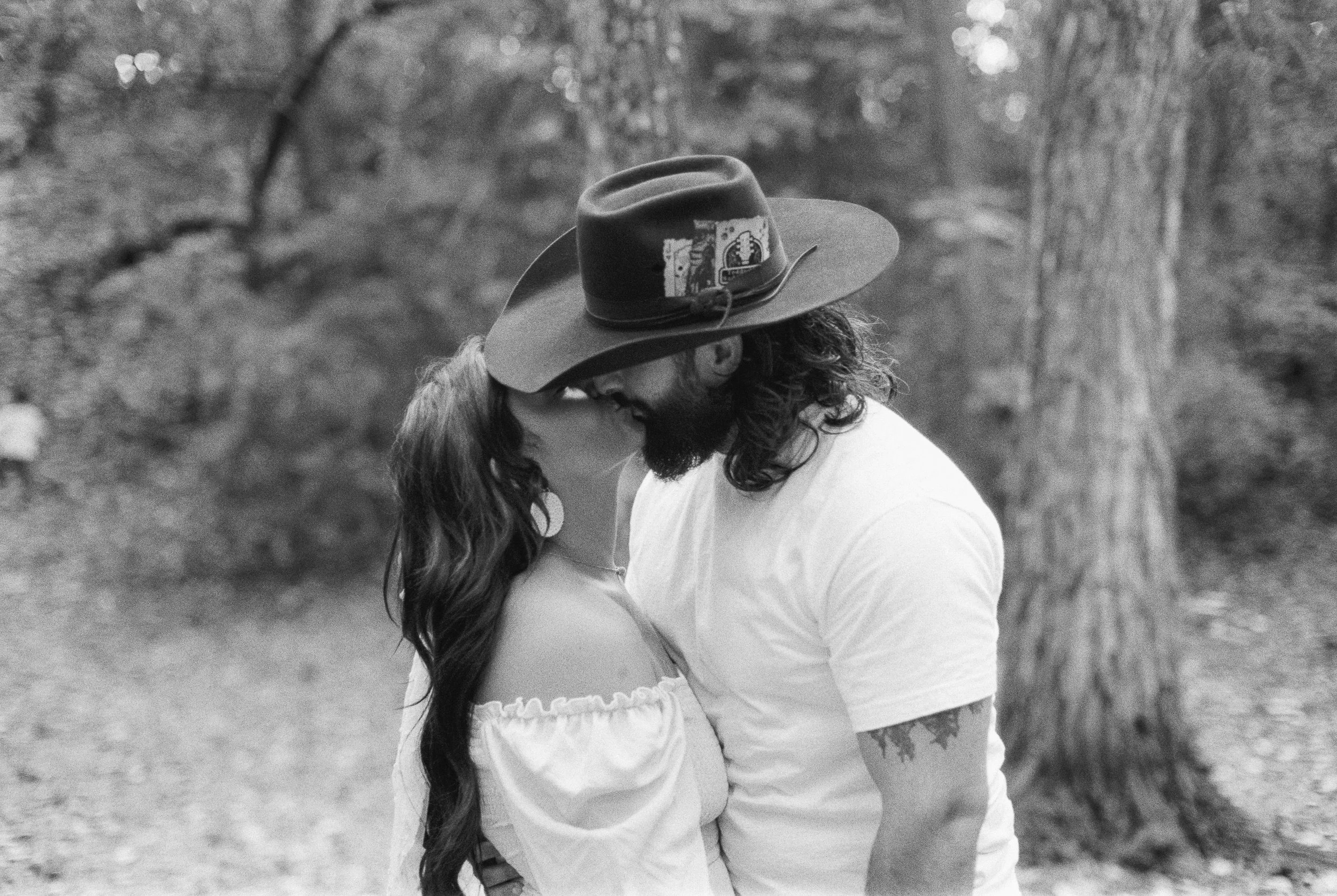 A black-and-white photo of a couple in an outdoor setting, wearing hats, with their foreheads touching and eyes closed, surrounded by trees and natural scenery.