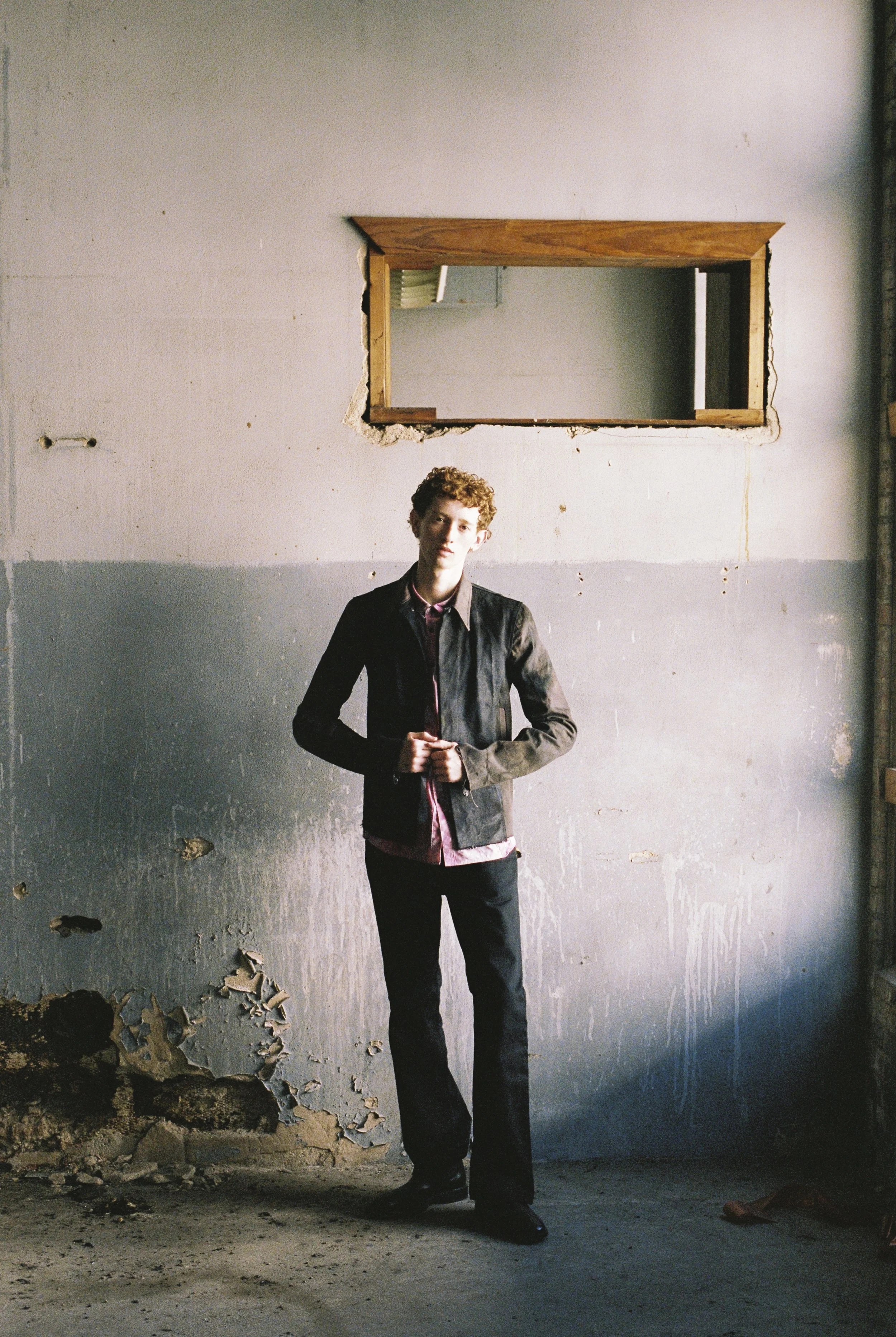 A young man with curly hair dressed in a leather jacket, red shirt, and black pants stands in a dilapidated room with peeling paint and exposed walls. He is looking at the camera with a neutral expression, with his hands clasped in front of him.