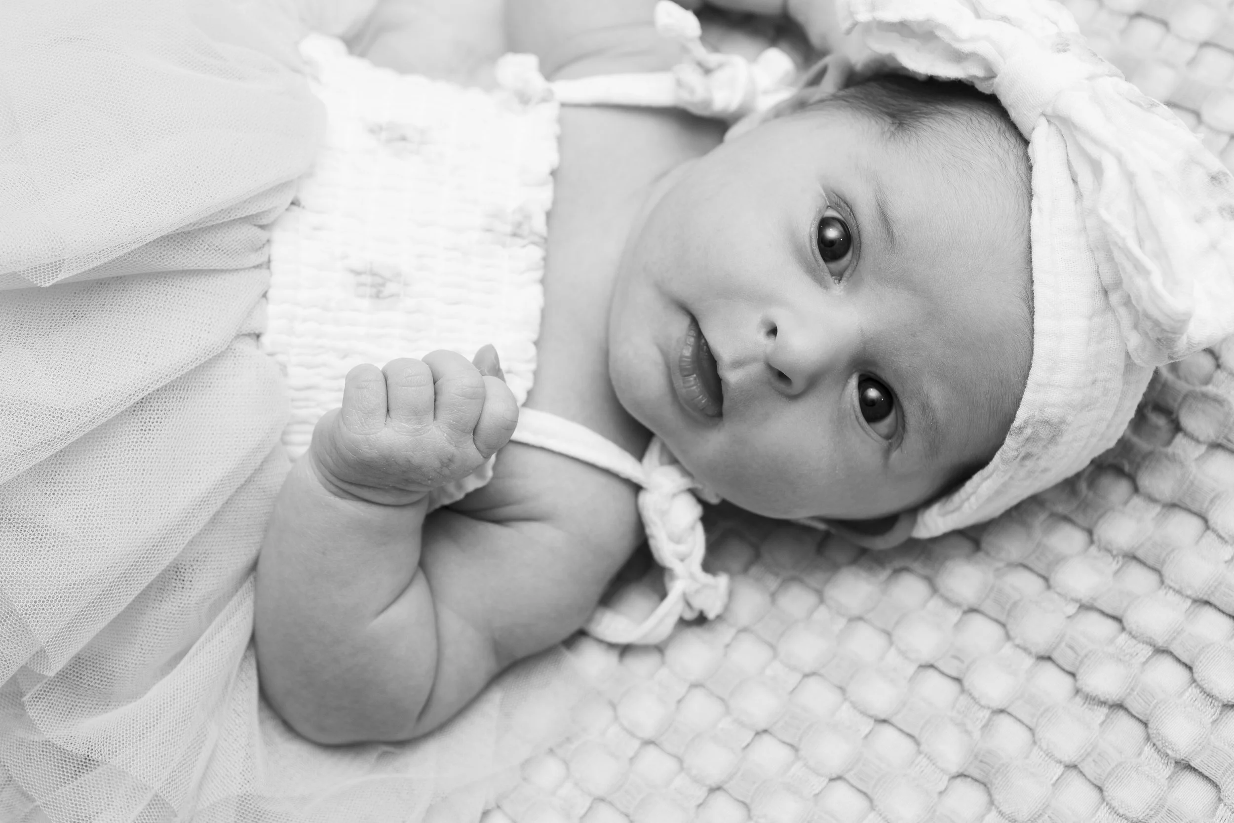 A baby lying on a textured blanket, wearing a headscarf and a sleeveless top, looking up with open eyes and a slight smile, holding one hand close to the face in black and white.