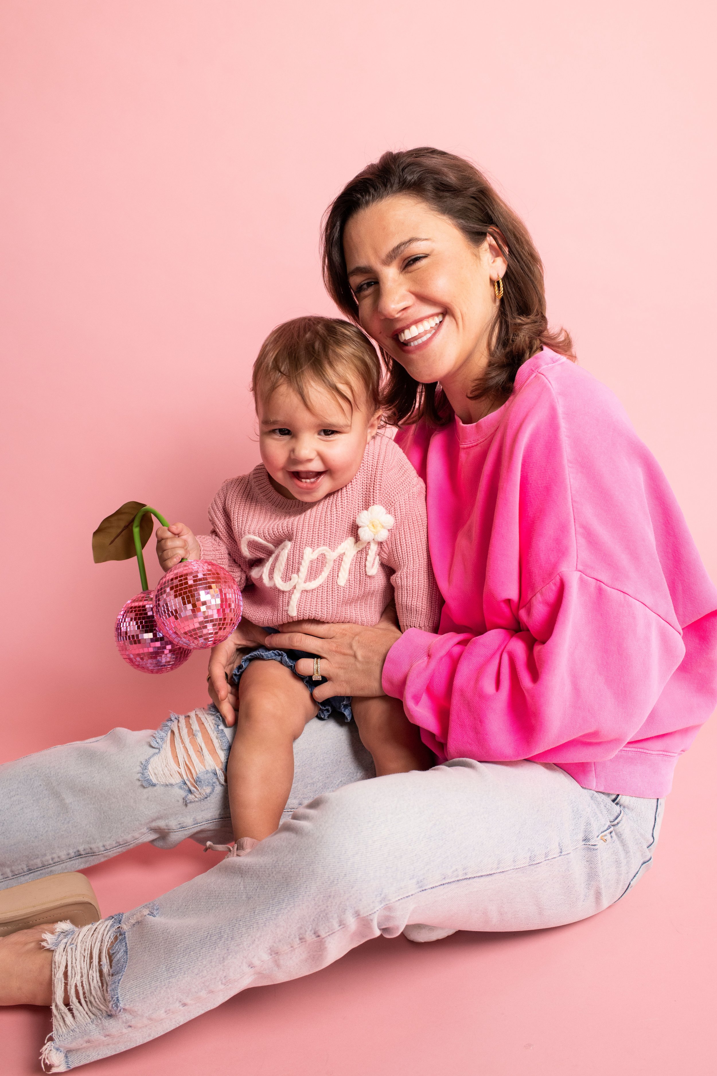 A woman and a young girl sitting against a pink background, smiling and holding a decorative cherry-shaped mirror.