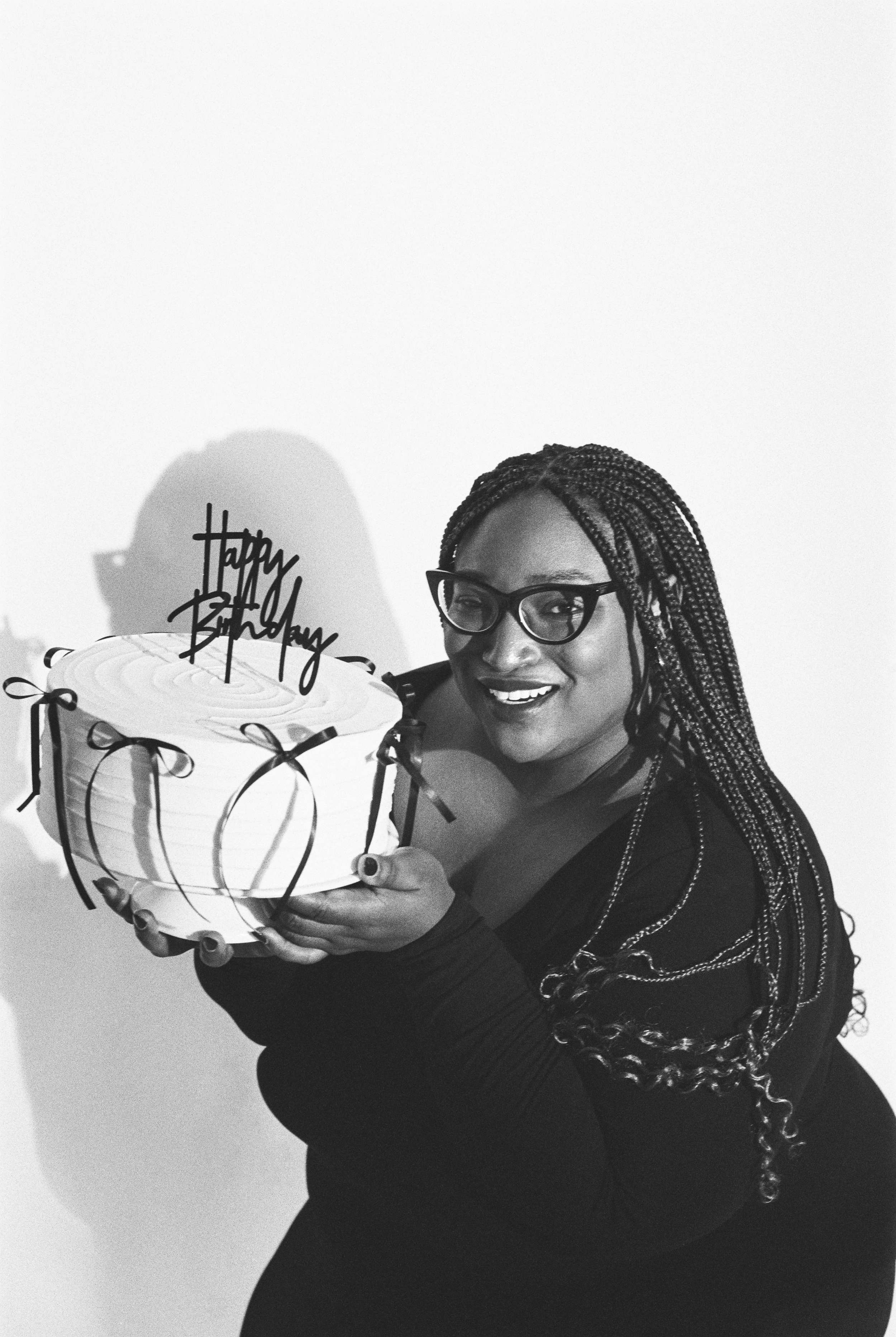 A woman with glasses smiling and holding a birthday cake with a "Happy Birthday" topper