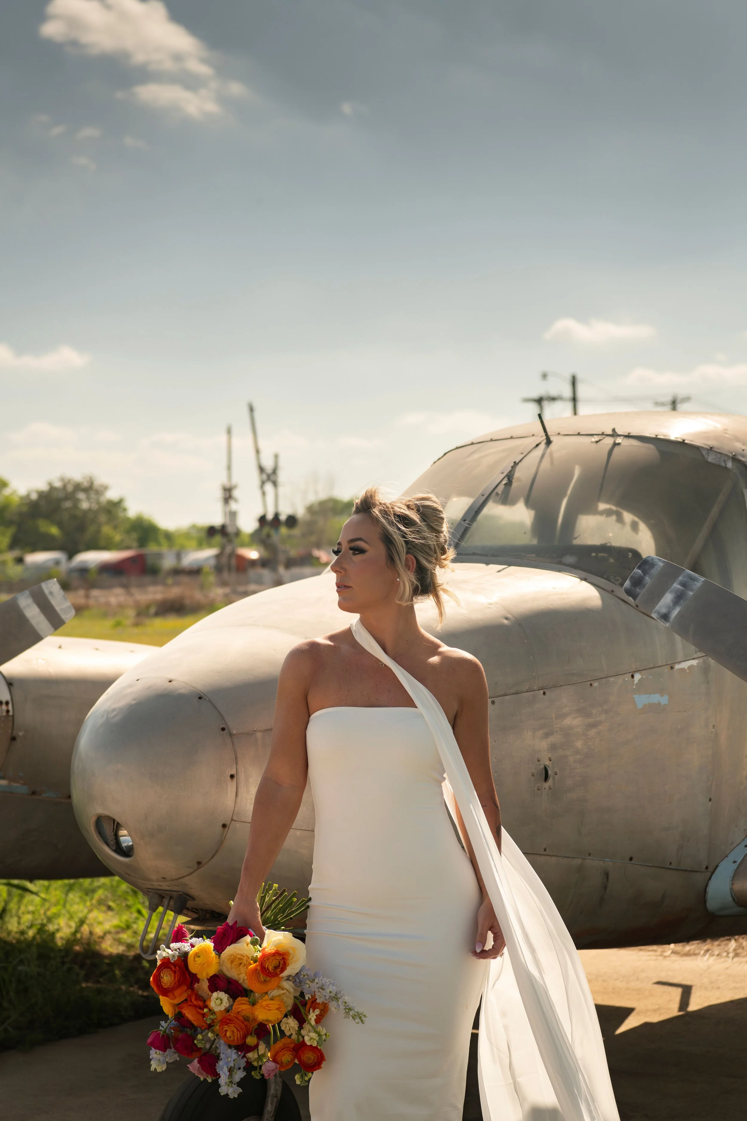 A woman in a white dress holding a bouquet of colorful flowers, standing in front of a vintage aircraft on a sunny day.