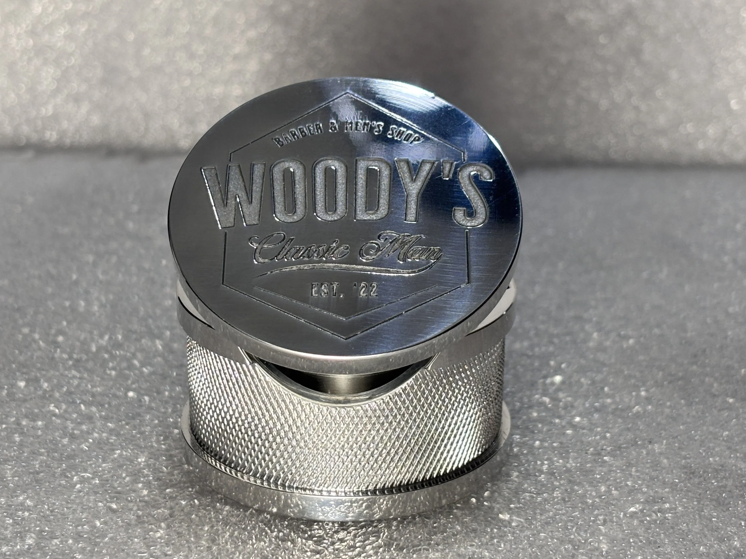 A shiny, metallic, circular metal cap with the inscription 'Woody's Classic Mach' and 'Est. '22' on it, placed on a textured surface.