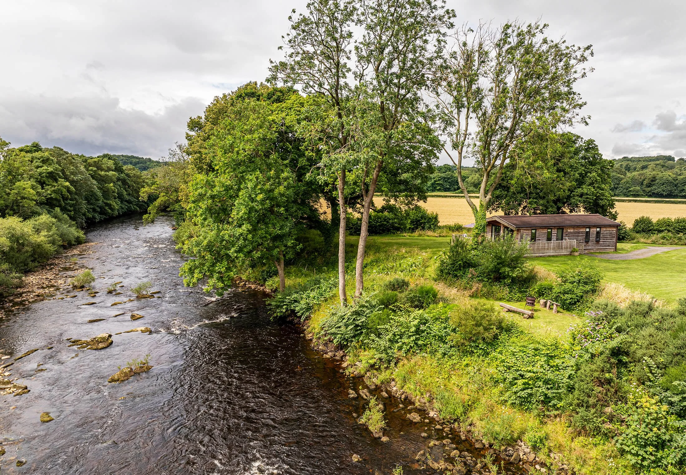 A riverside landscape with a wooden cabin surrounded by tall trees and lush greenery. The flowing river on the left has rocks and the area is bordered by fields with a cloudy sky overhead.