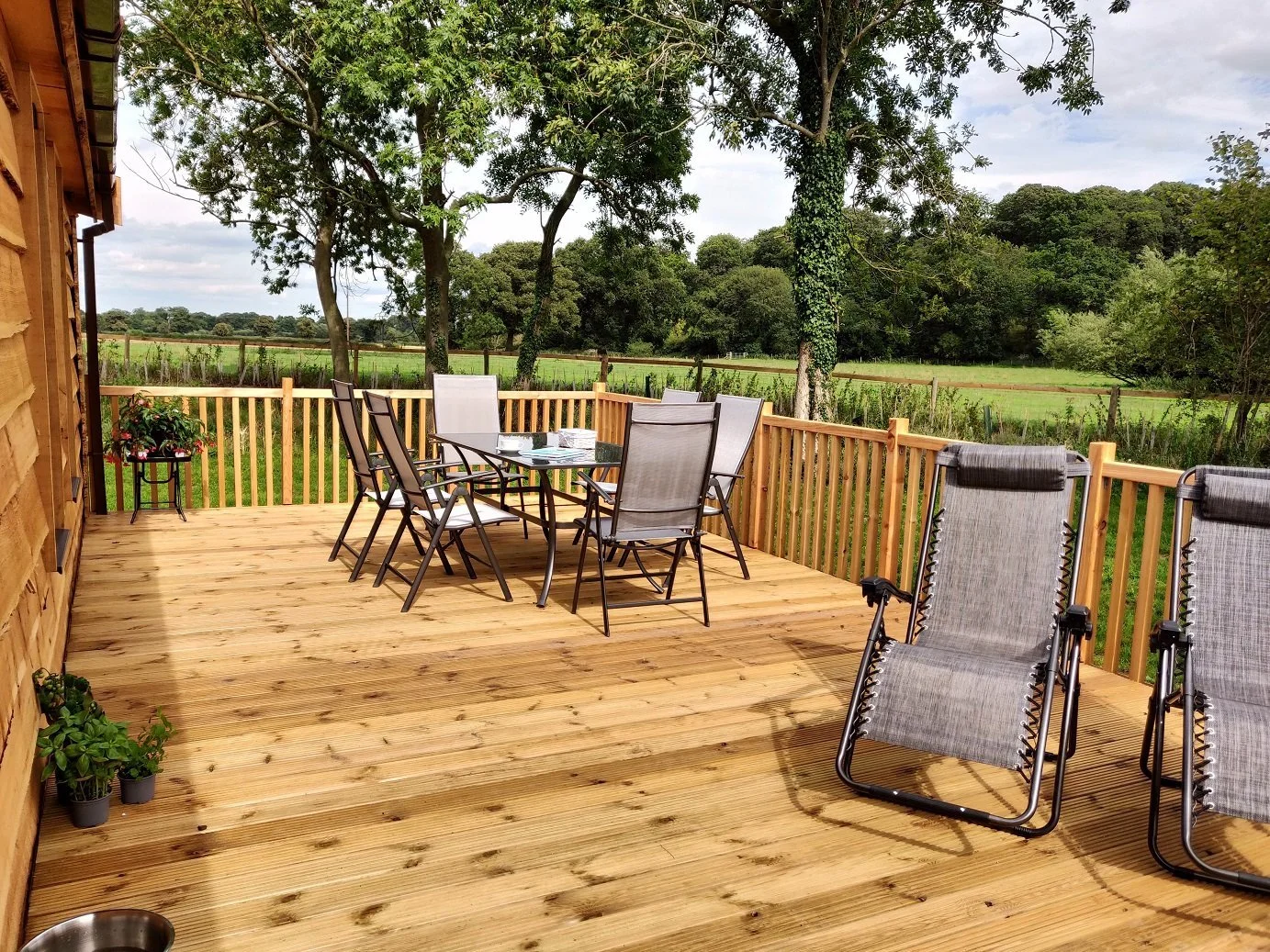 A Riverdale Yorkshire Luxury Lodge outdoor decking with a table and six chairs, two lounge chairs, and potted plants, overlooking green fields and trees under a partly cloudy sky.