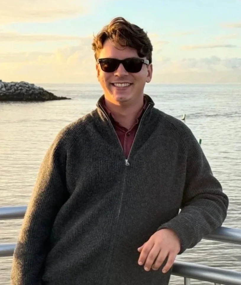A young man with short brown hair wearing sunglasses and a dark zip-up sweater, smiling while standing by a metal railing near the water at sunset.