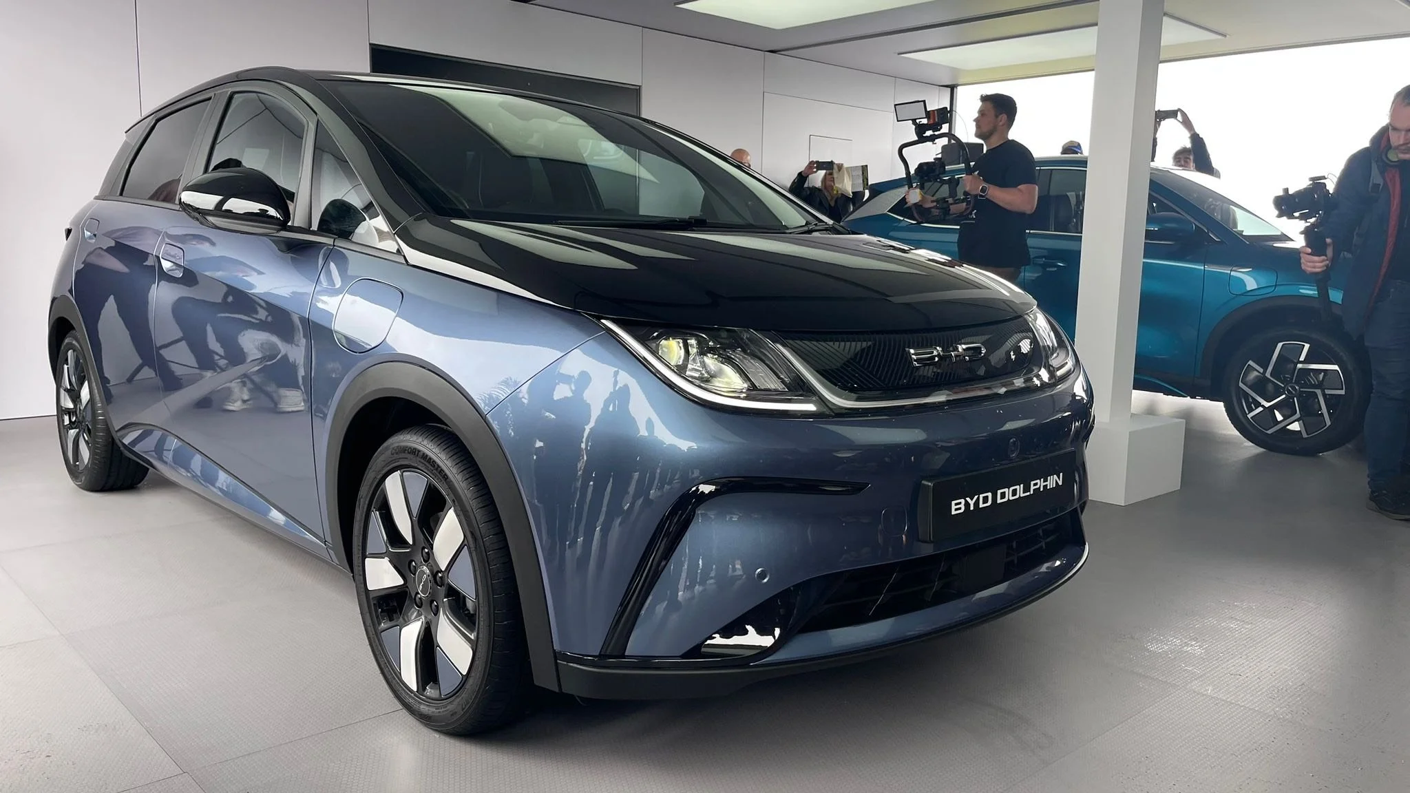 Blue and black BYD Dolphin electric car displayed at an indoor event with cameras and attendees taking photos.