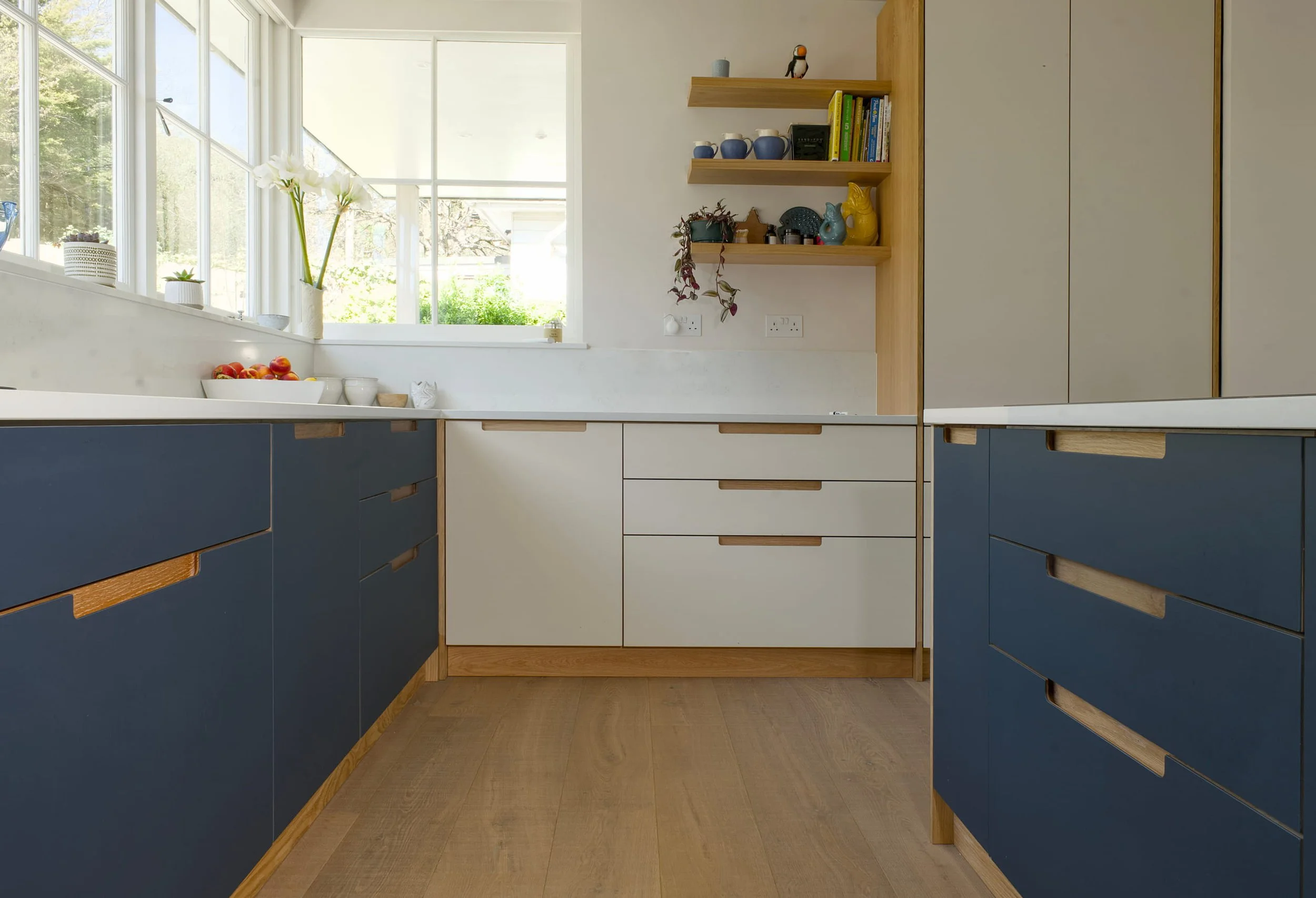 U-shape view down the bespoke kitchen showing smokey blue base units, open American white oak shelves, and wide-plank oak flooring