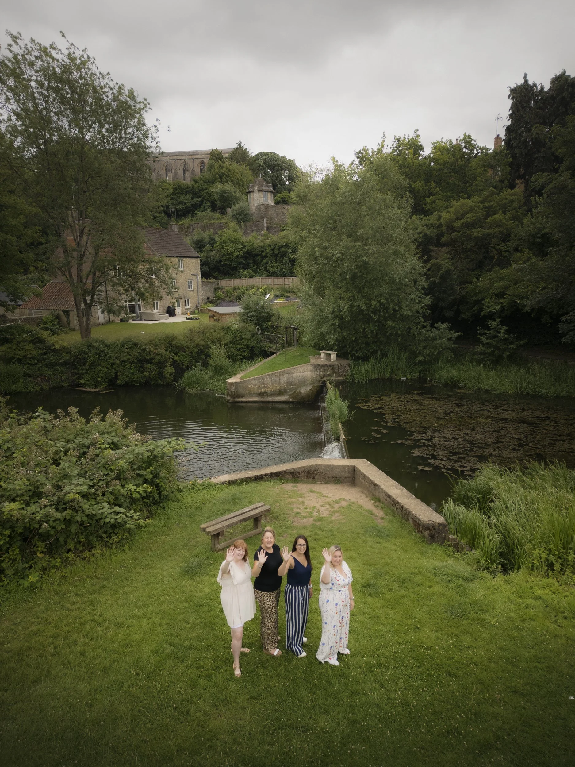 Four women standing on grass near a river, waving at the camera, with trees, a stone house, and a hillside with a church in the background under an overcast sky.