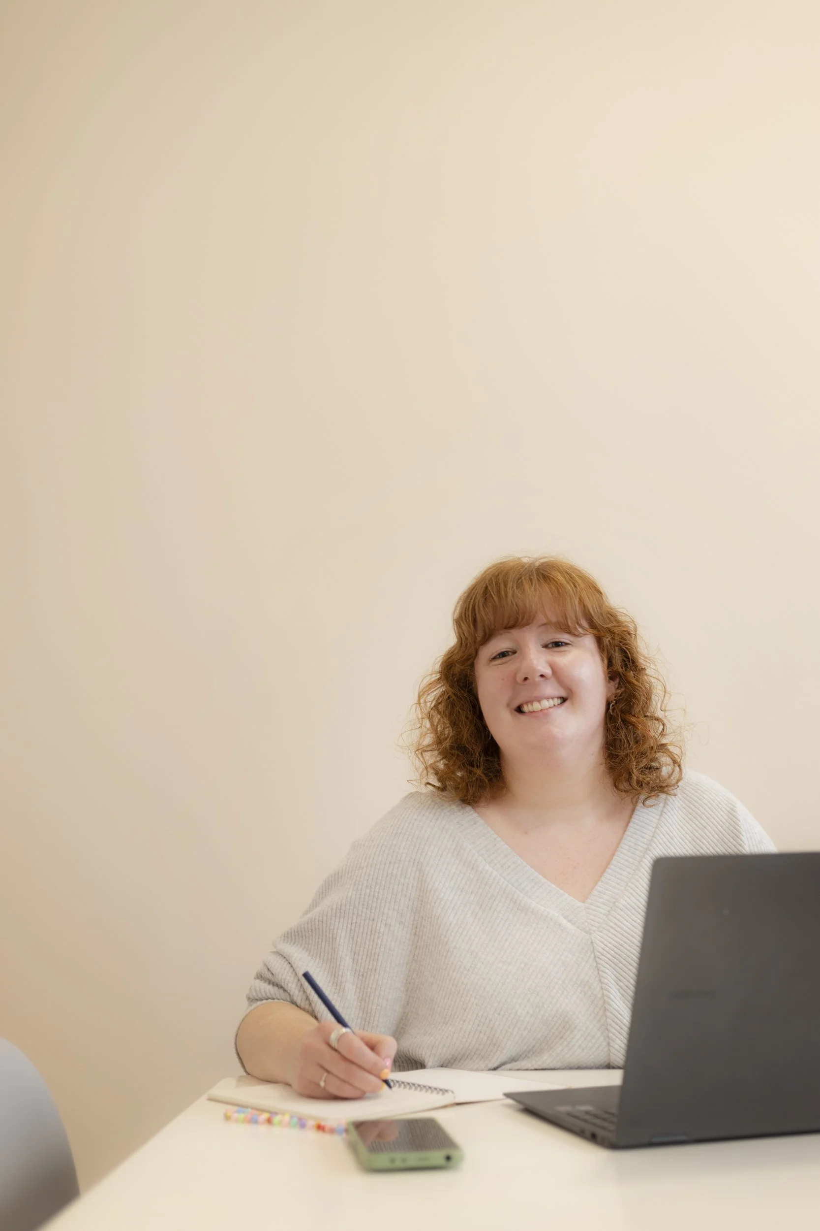 A woman with ginger hair sit at a desk with a laptop in front of her. She grins at the camera