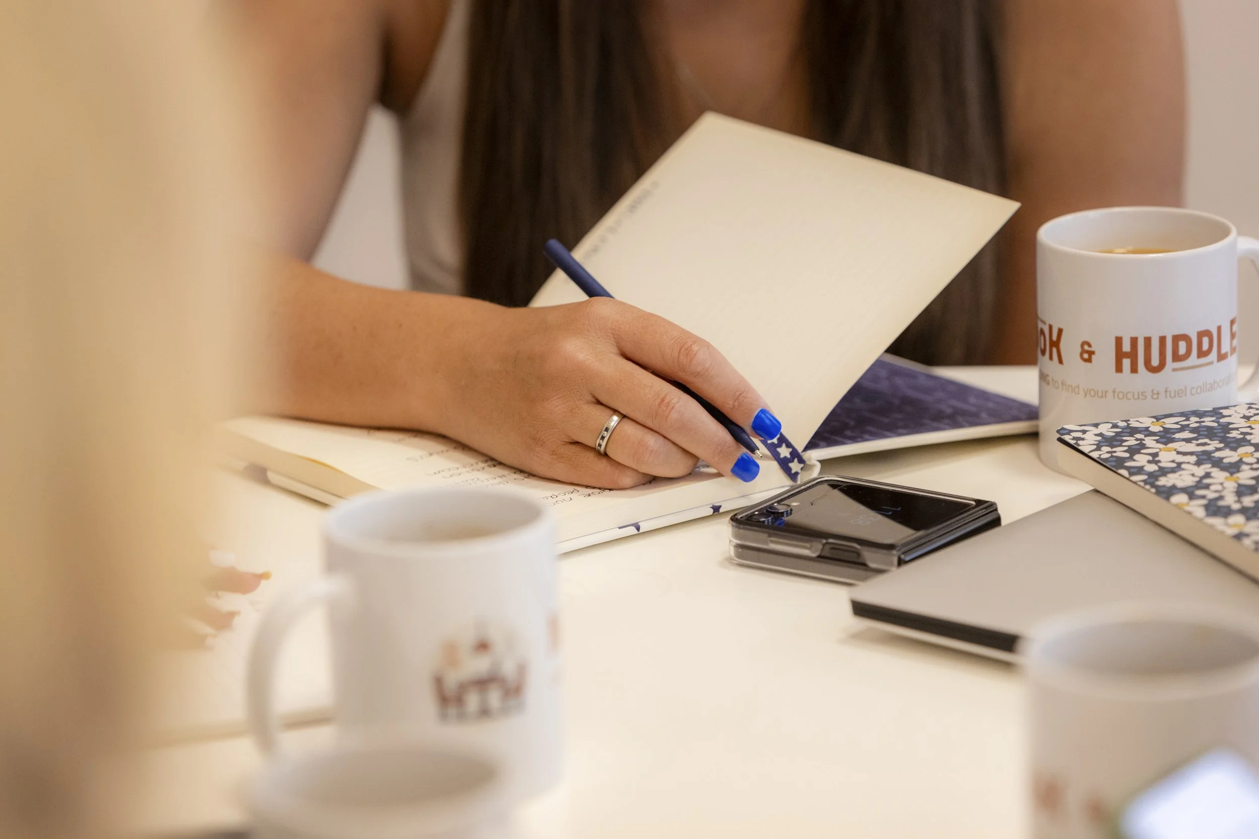 A lady with blue painted fingernails writes in a notebook which is on a desk with a smartphone and mug.