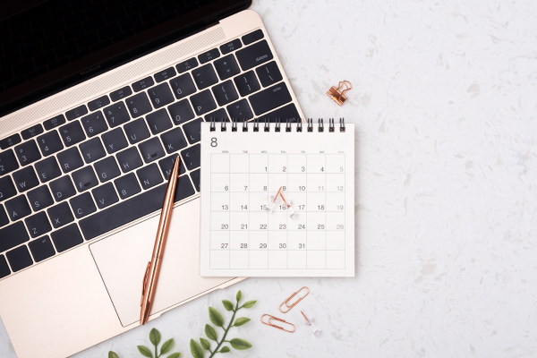 A desk with a laptop, a gold pen, a small desktop calendar showing August 23, and some office supplies including paper clips and a binder clip, on a white surface with green leaves.