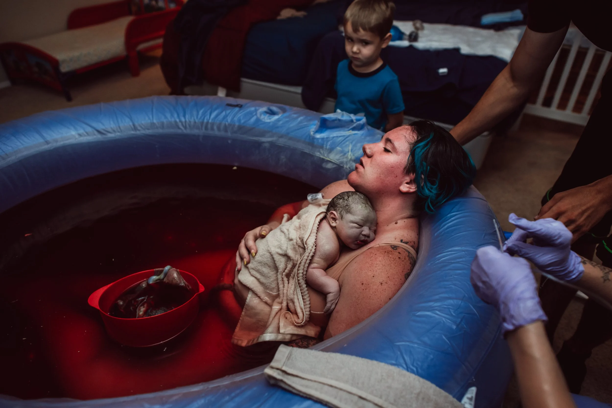 Woman giving birth in a water birth tub with a newborn baby resting on her chest, while a boy watches in a bedroom.