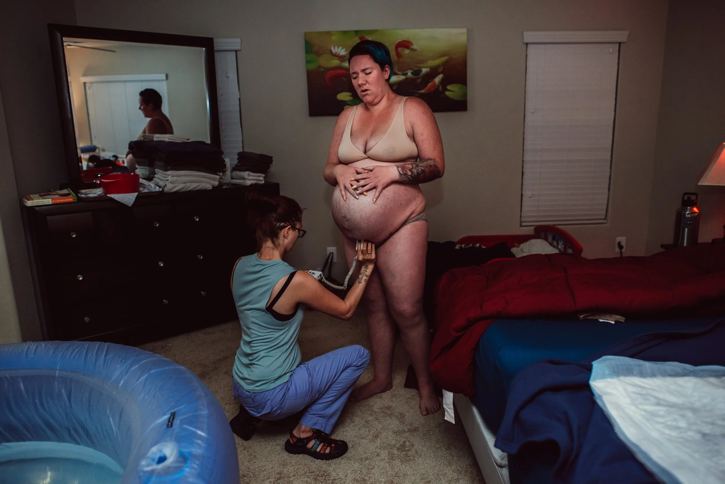 A woman with tattoos and short hair, wearing a beige bra and matching underwear, is standing in a bedroom, holding her pregnant belly with both hands, eyes closed. A nurse kneeling in front of her is taking her blood pressure with a cuff on her arm. The room has a large mirror, a dresser with clothes on top, a bed with red and blue bedding, a window with closed blinds, and a colorful painting of water lilies and swans on the wall.