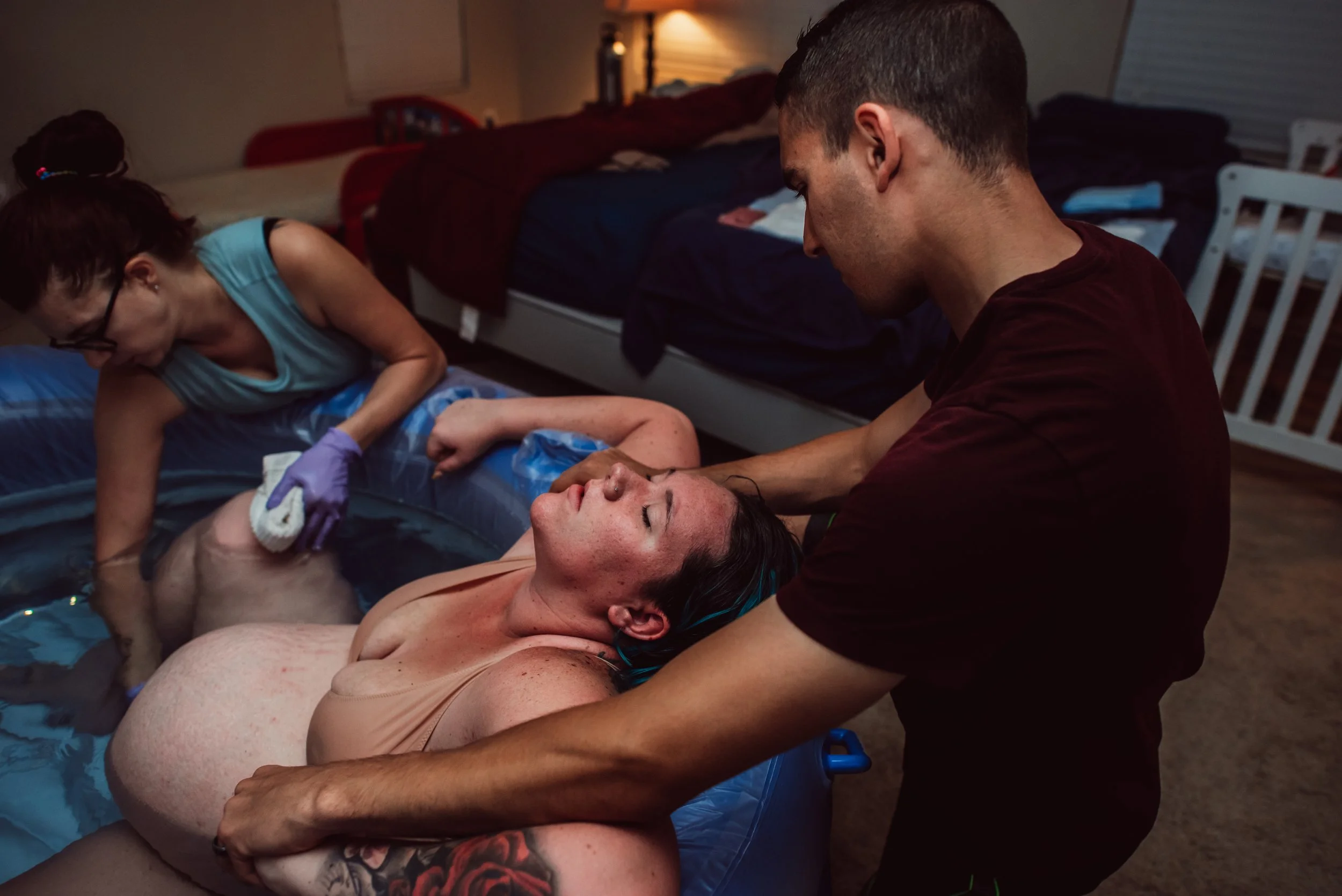 Woman giving birth in a birthing pool at home with a man supporting her, a woman assisting with the delivery, in a warmly lit room with beds and a crib in the background.