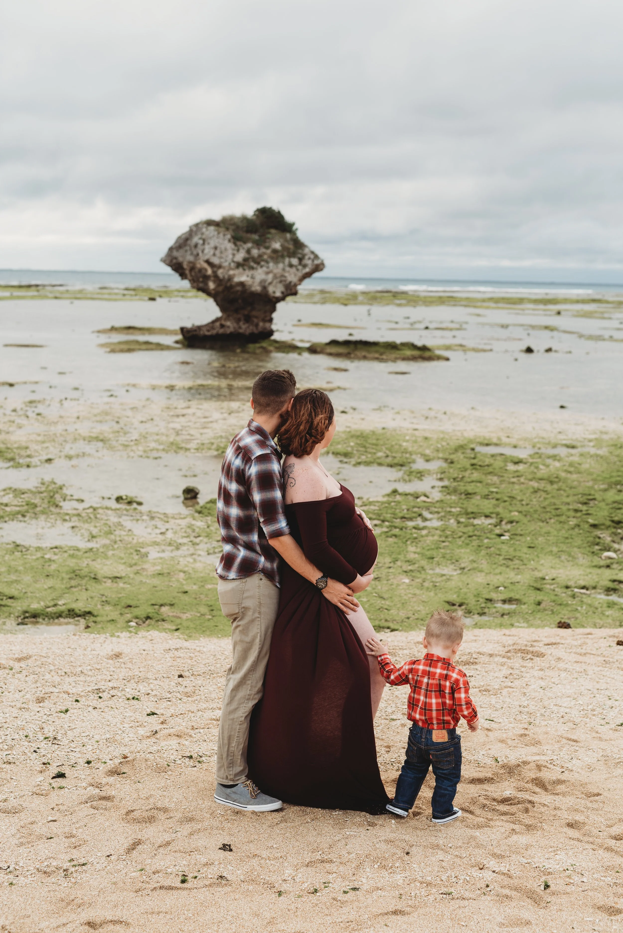 A pregnant woman and a man are standing on a beach with a young boy. The man is holding her belly while she is touching it. The boy is holding her hand. In the background, there is a rock formation on the beach and the sea with cloudy skies overhead.