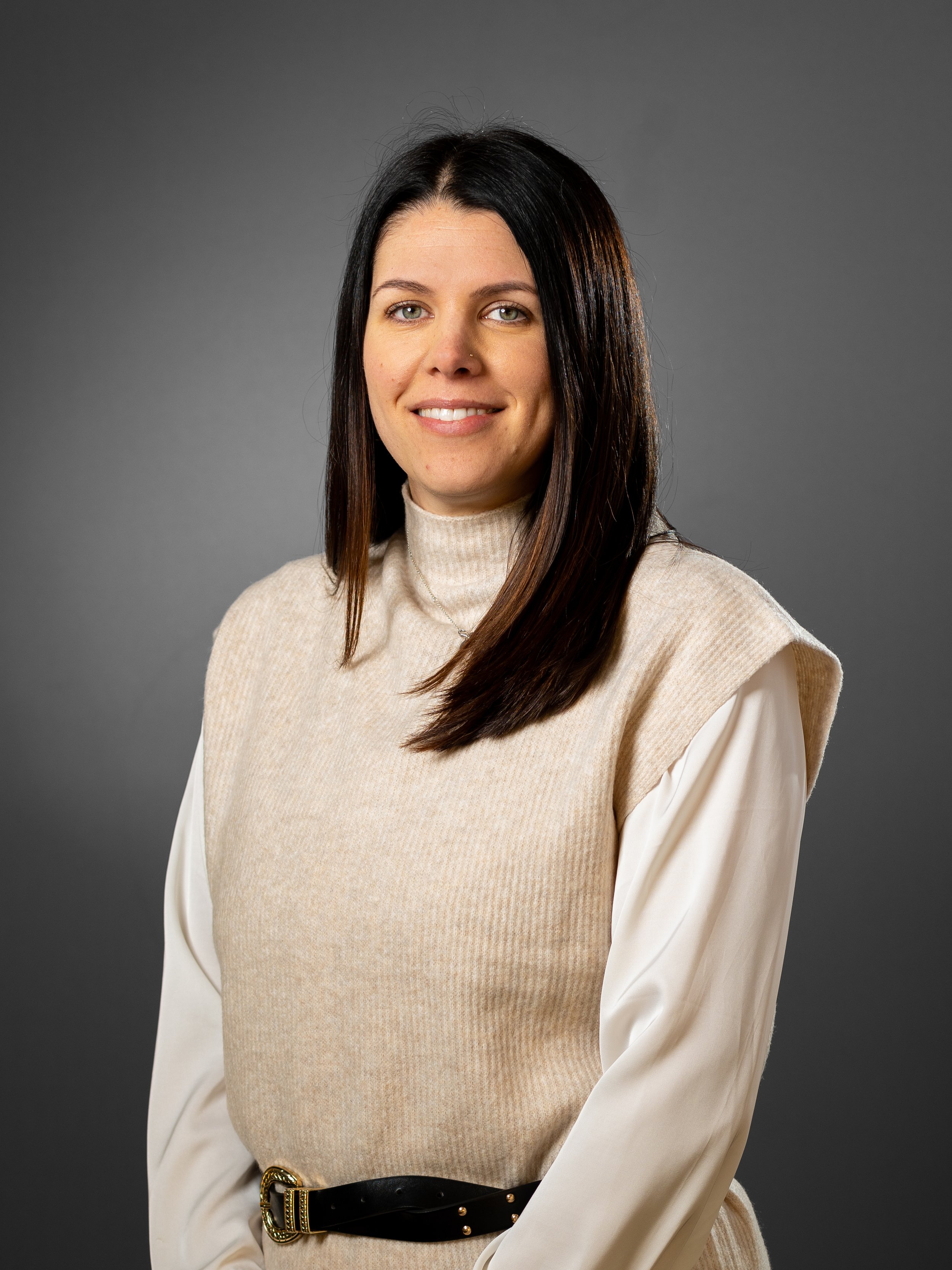 Woman with long dark hair wearing a beige sleeveless sweater over a white shirt, smiling against a gray background.