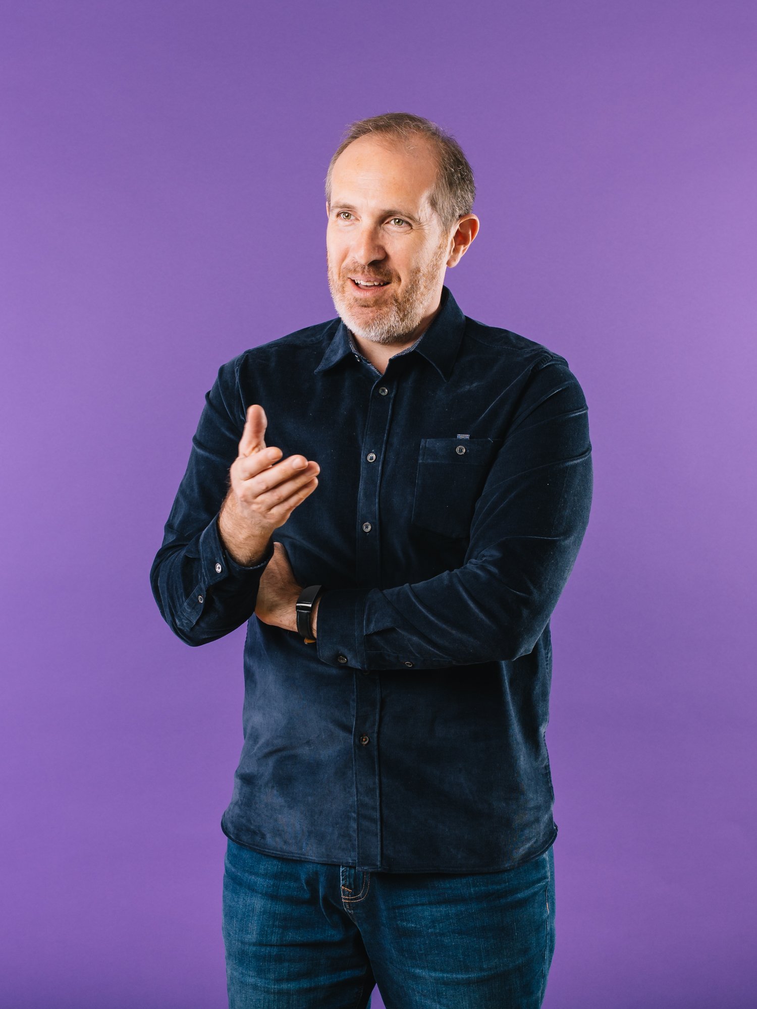 Man in a dark shirt gesturing against a purple background.