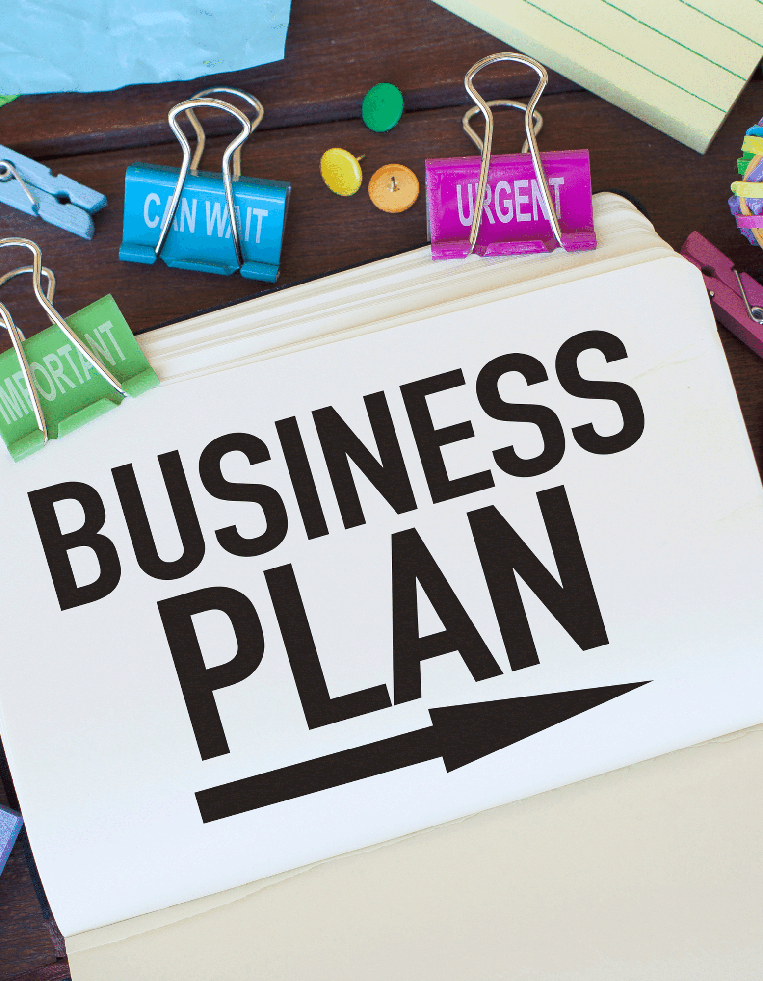 A planner with the words "BUSINESS PLAN" and an arrow on the cover, surrounded by colorful paper clips and thumbtacks labeled 'Important,' 'Can Wait,' and 'Urgent' on a wooden desk.