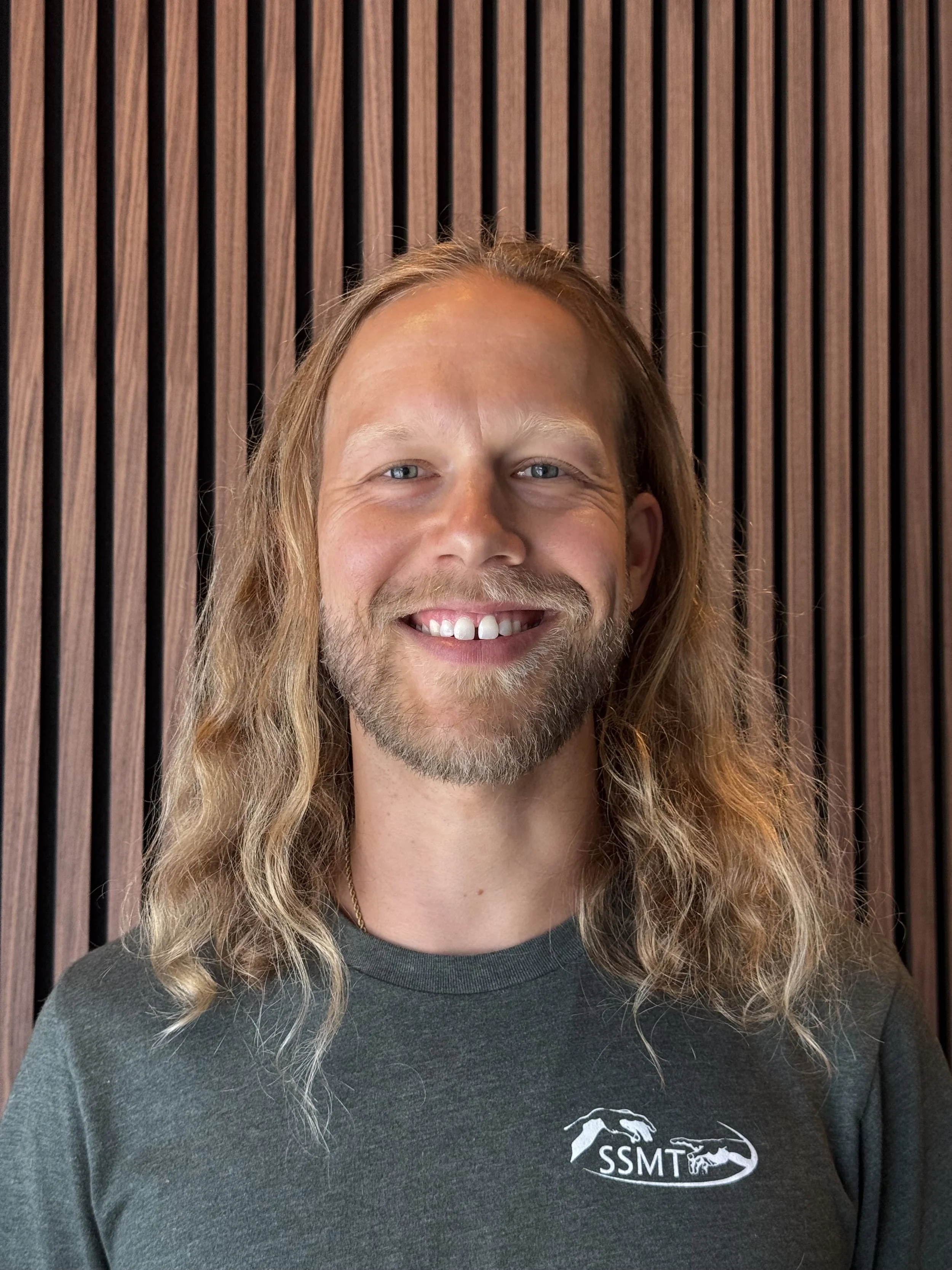 A man Christopher Ostema with a beard and a bandana on his head smiling in a dense jungle, holding a yellow fruit in one hand and a long pole or machete in the other. BioLIT spa licensed Massage Therapist