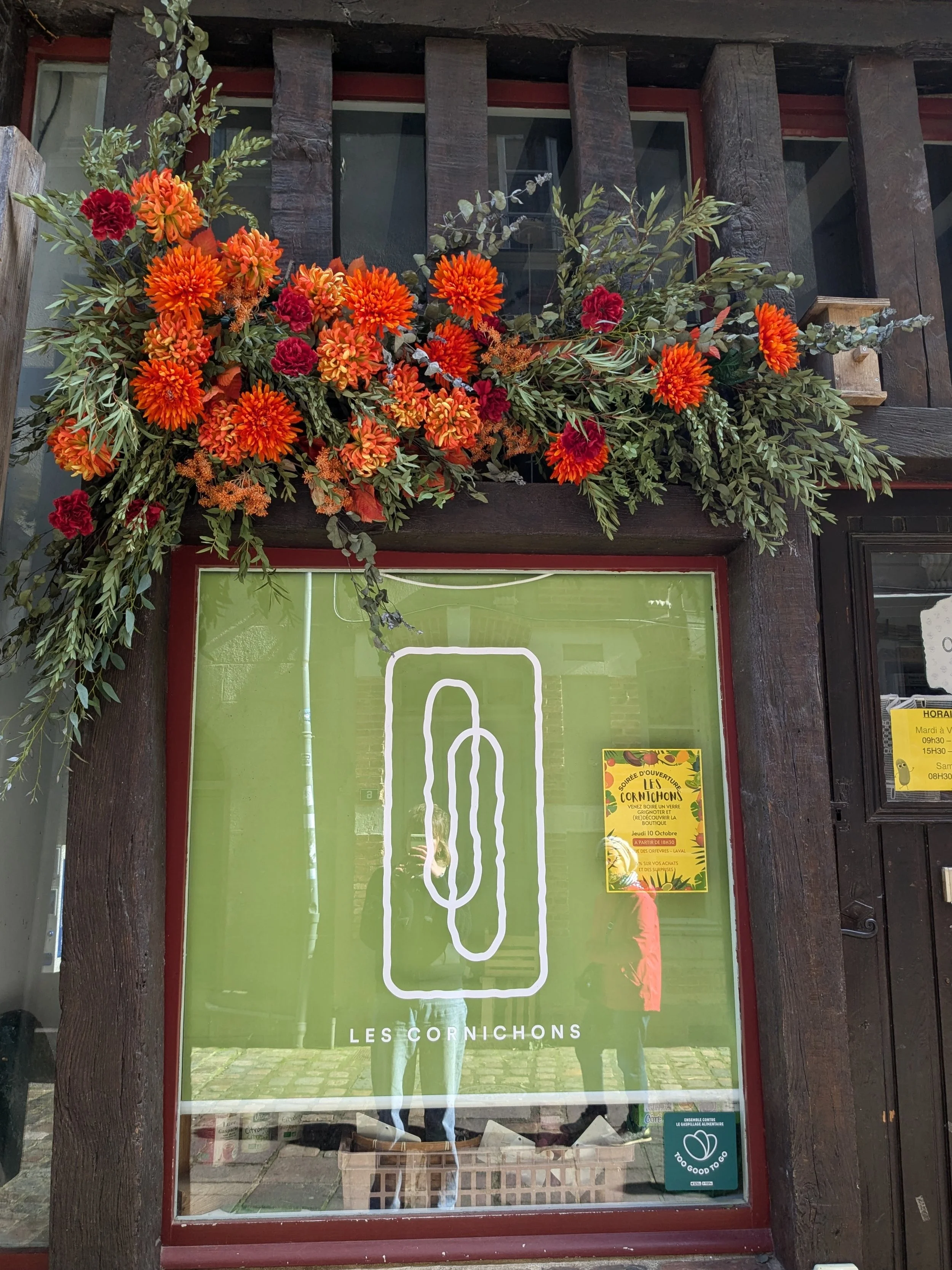 Vitrine avec un panneau vert,logo blanc représentant une silhouette de cornichon, texte 'Les Cornichons'. Décorations florales avec des fleurs orange et rouges en haut de la vitrine. Un autocollant en bas à droite indique qu'il est 'Too Good To Go'.