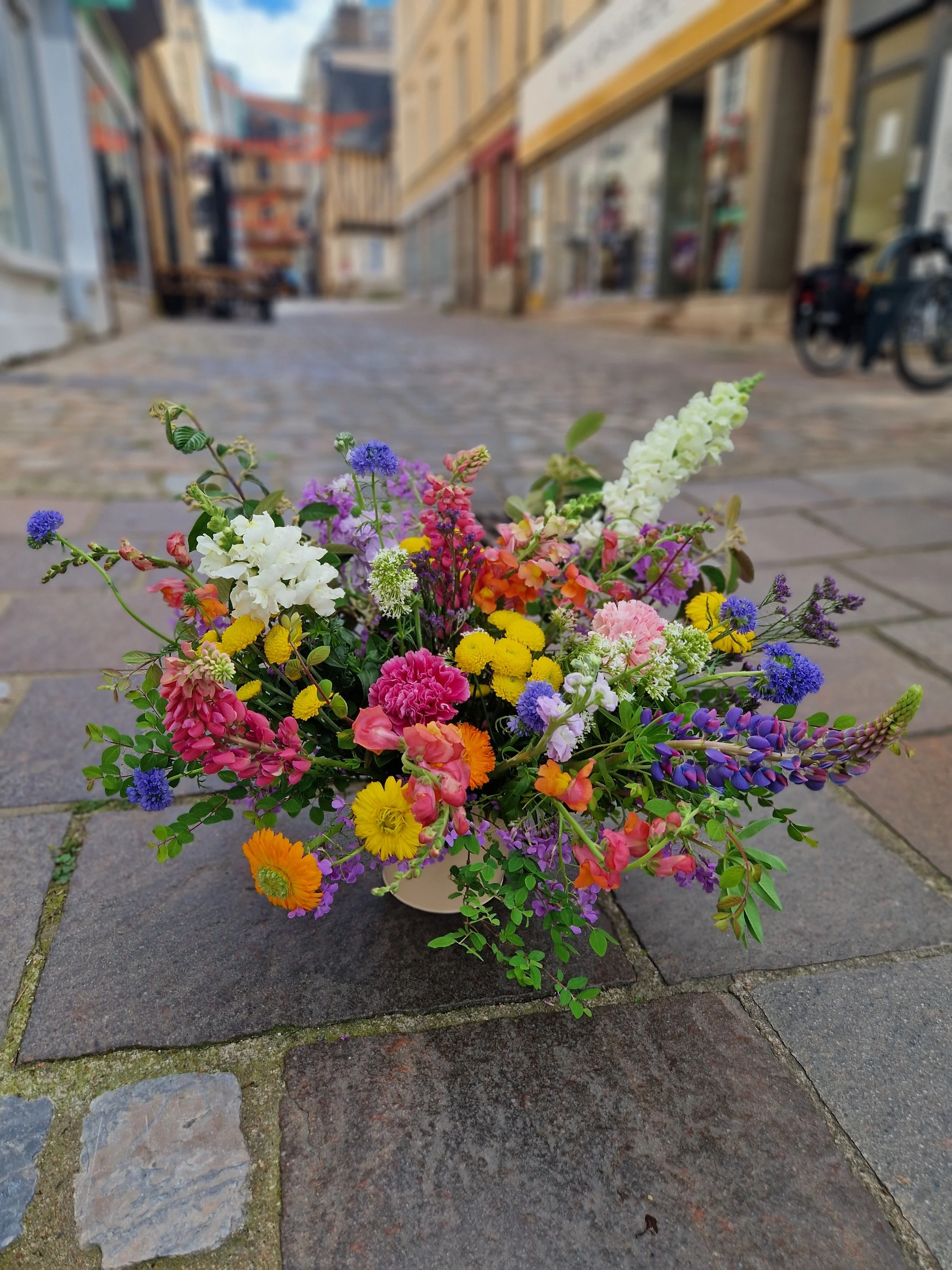 Un bouquet de fleurs colorées comprenant des fleurs blanches, violettes, roses, jaunes et orange, placé sur un pavé de rue.