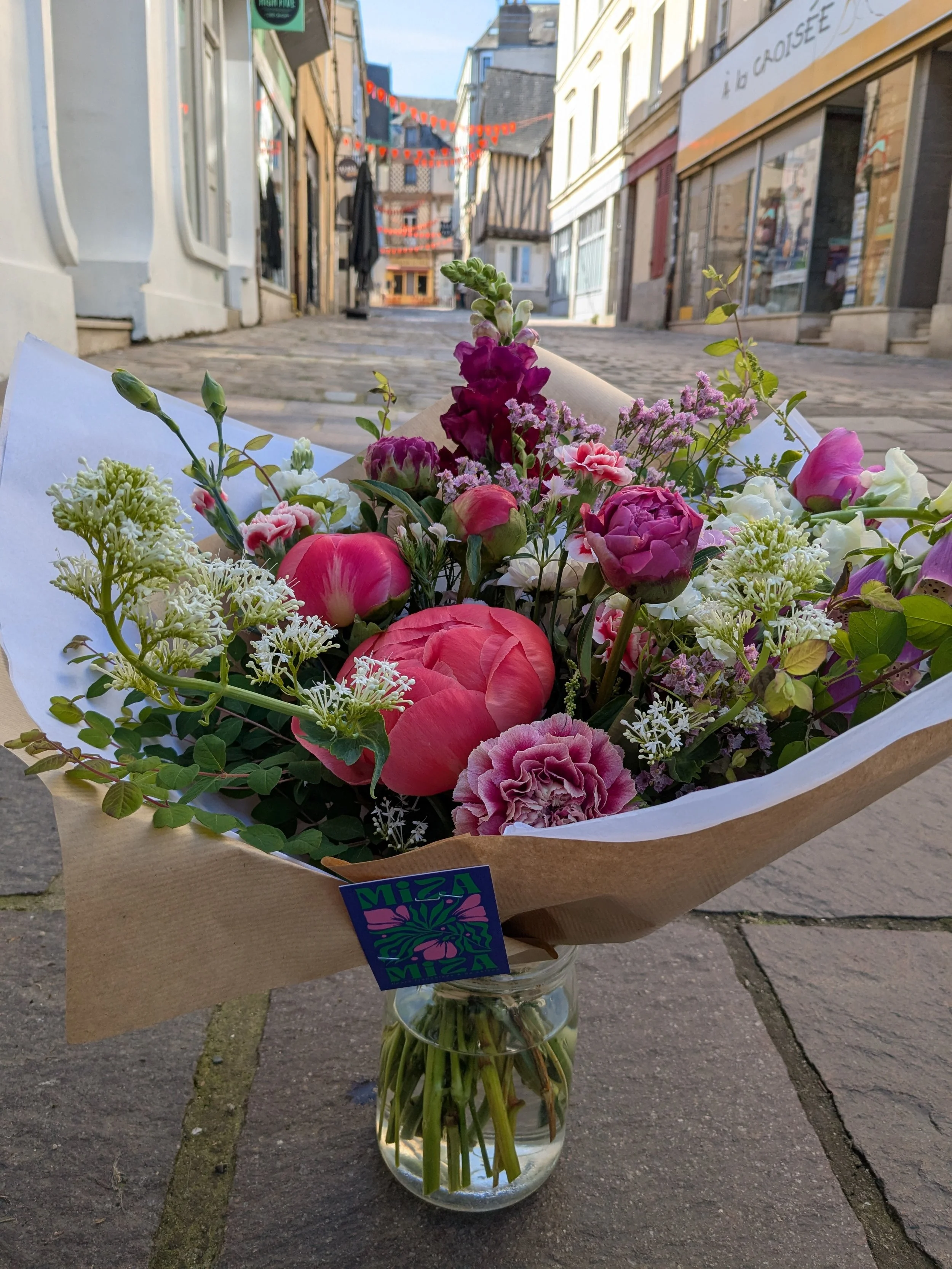 Un bouquet de fleurs colorées dans un vase en verre placé sur un trottoir de rue. Des bâtiments anciens bordent la rue, décorée de guirlandes lumineuses rouges suspendues en suspension.