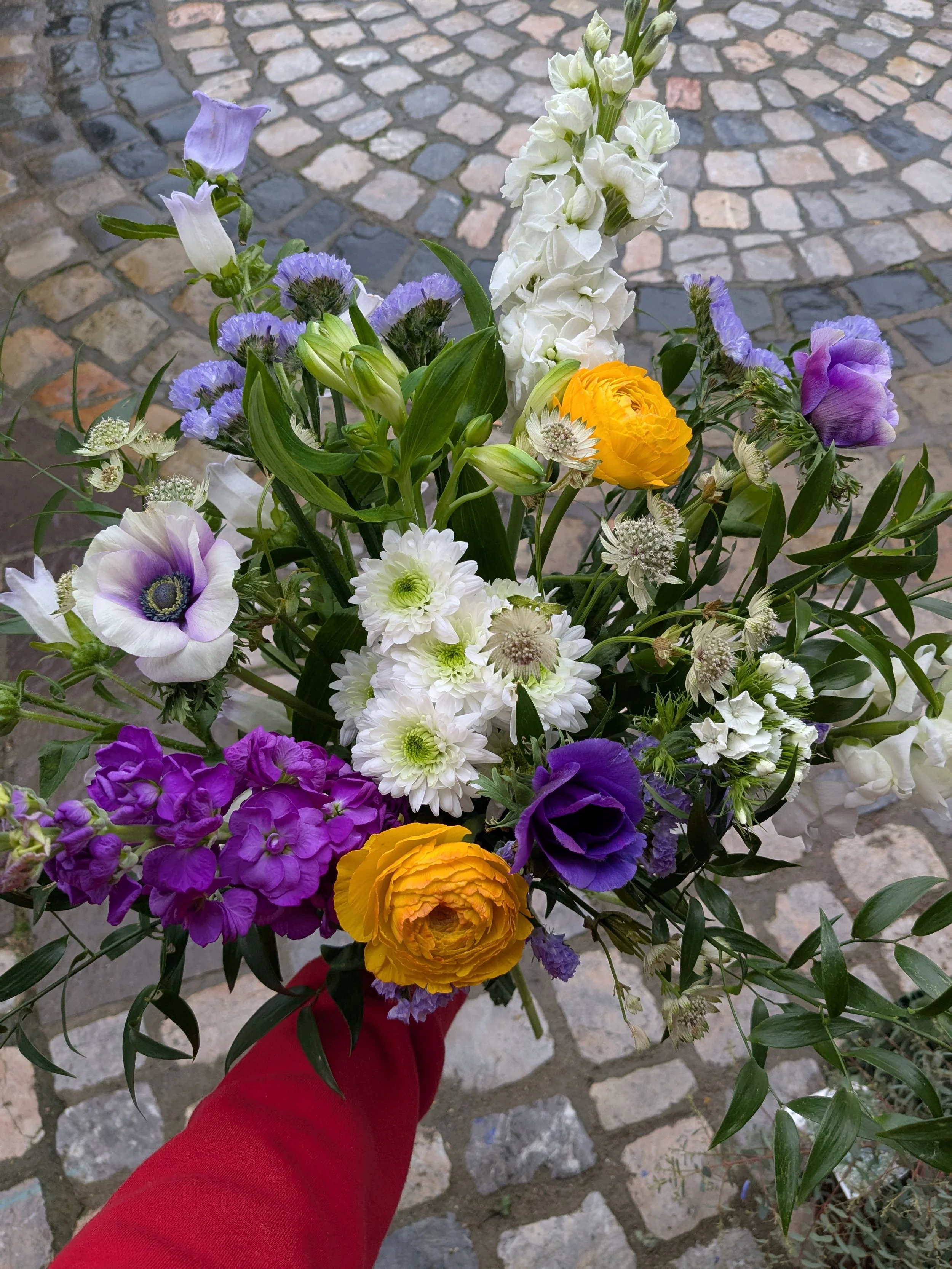 Un bouquet de fleurs colorées avec des roses jaunes, des lis blancs, des chrysanthèmes blancs, des œillets violets et des fleurs violettes, tenu par une personne portant un vêtement rouge sur un sol en pavés.