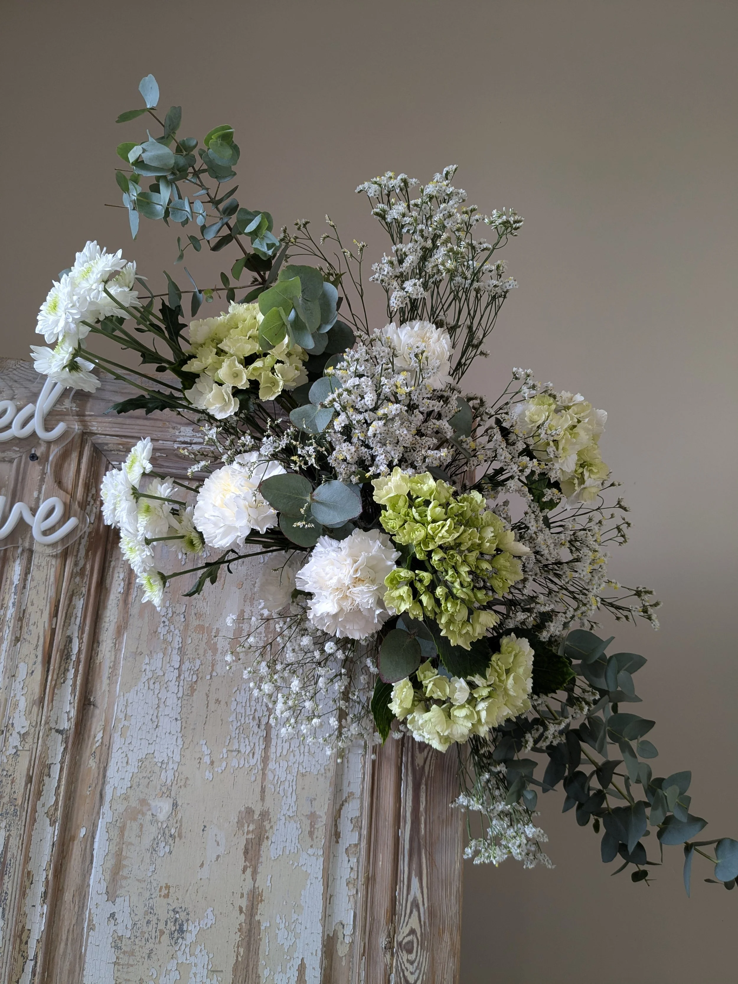 Composition florale avec des fleurs blanches et vertes, des feuilles d'eucalyptus et des petites fleurs blanches, posée sur une table en bois avec un fond neutre.