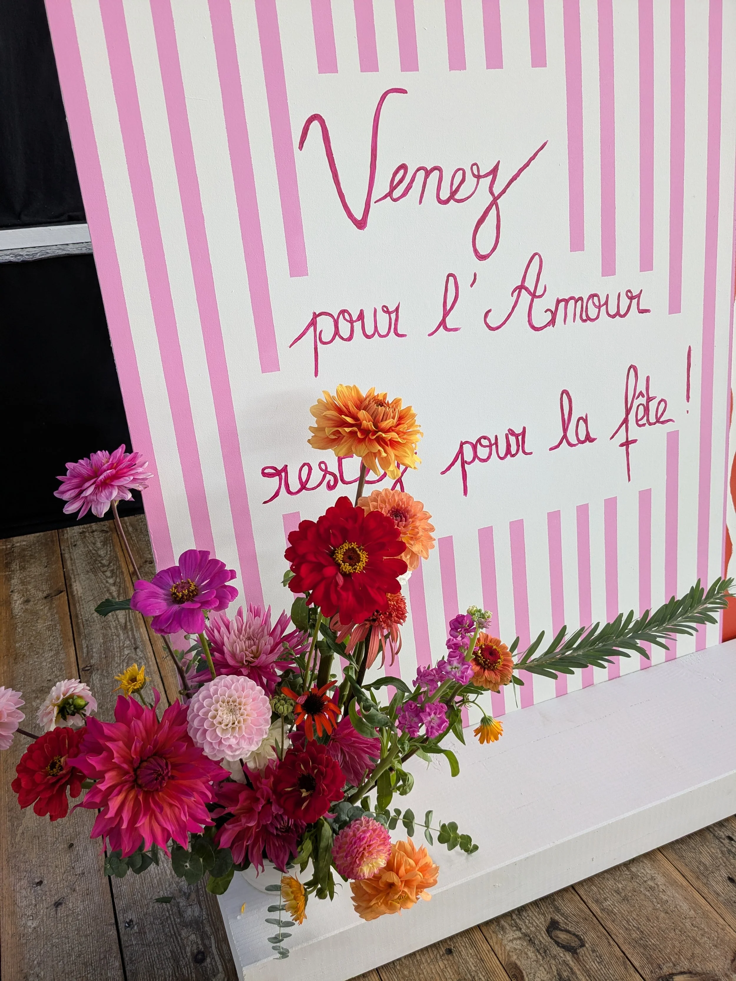 Un panneau blanc avec des rayures roses et du texte écrit à la main en rose, entouré de fleurs colorées, probablement pour une fête ou un événement. Le texte mentionne Venise, l'amour et la fête.