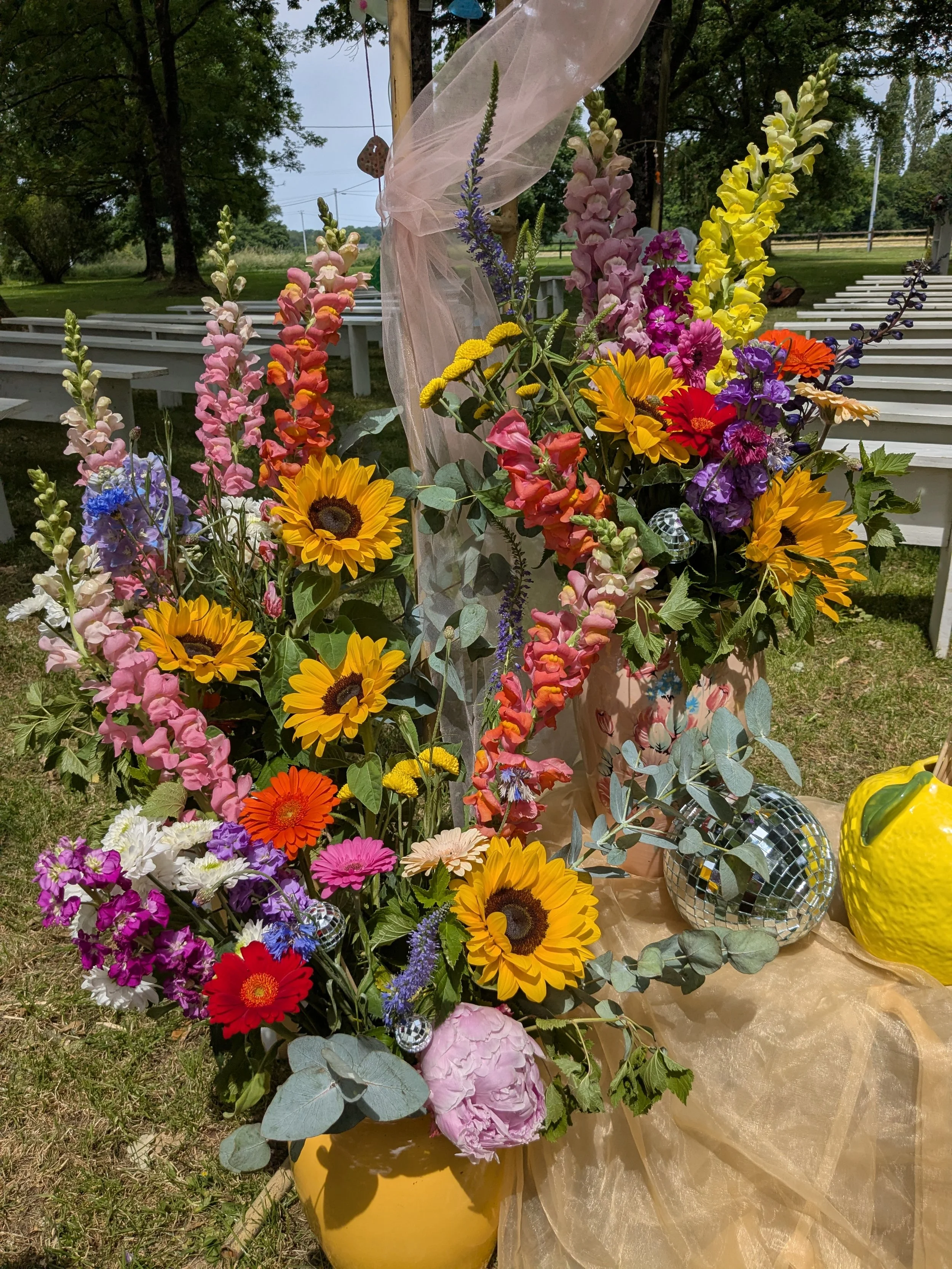 Un arrangement floral coloré comprenant des tournesols, des zinnias, des roses, des lys, des fleurs violettes et des feuillages verts, placé sur une surface en tissu doré à l'extérieur, avec des bancs en bois et des arbres en arrière-plan.