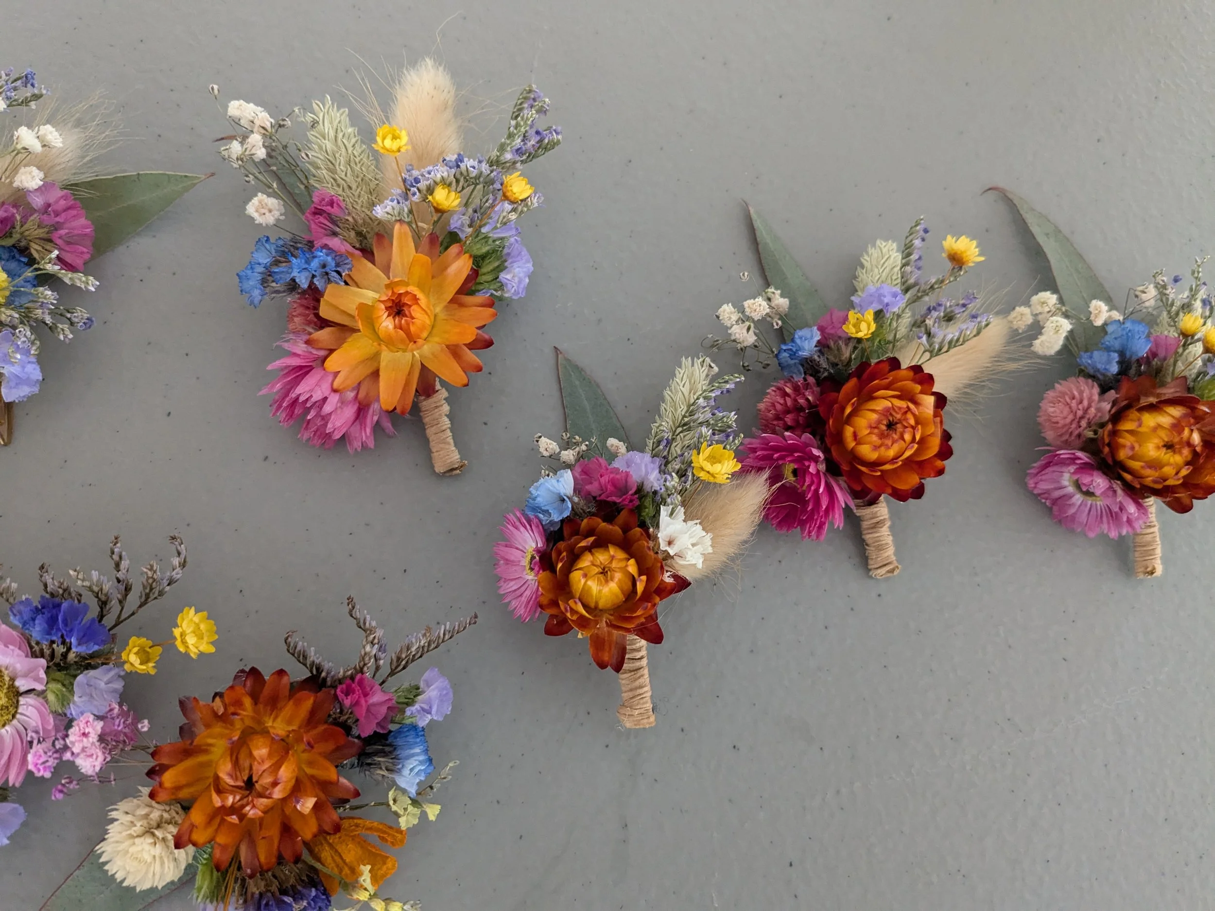 Composition de boutons de fleurs séchées colorées, incluant des fleurs orange, jaunes, roses, violettes, blanches et bleues, avec des feuilles vertes, certains enroulés avec de la ficelle