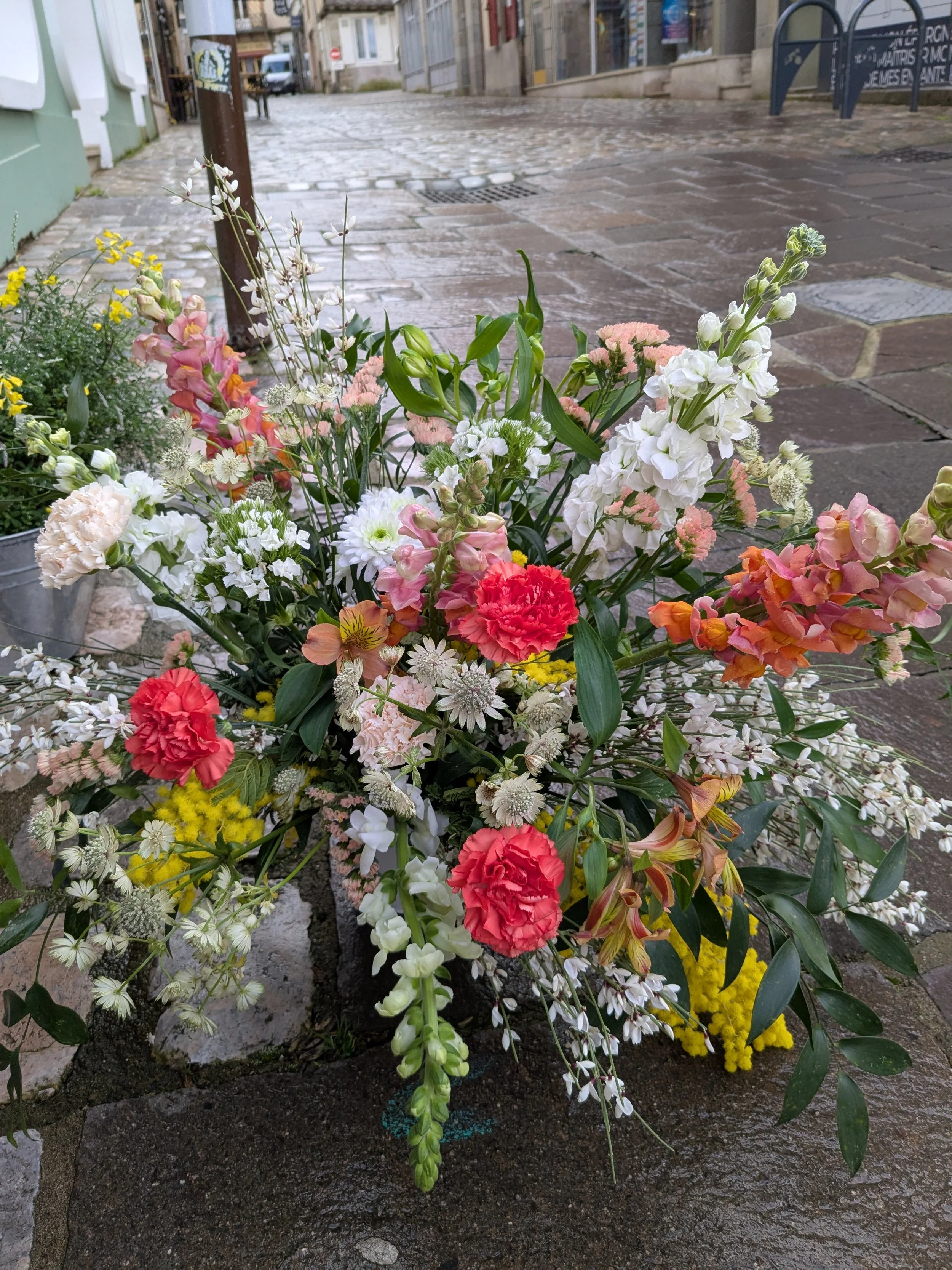 Un arrangement floral vertical avec des fleurs colorées, dont des carnations rouges, des fleurs blanches, des roses, des géraniums et d'autres fleurs, placé en ville sur un sol pavé humide.