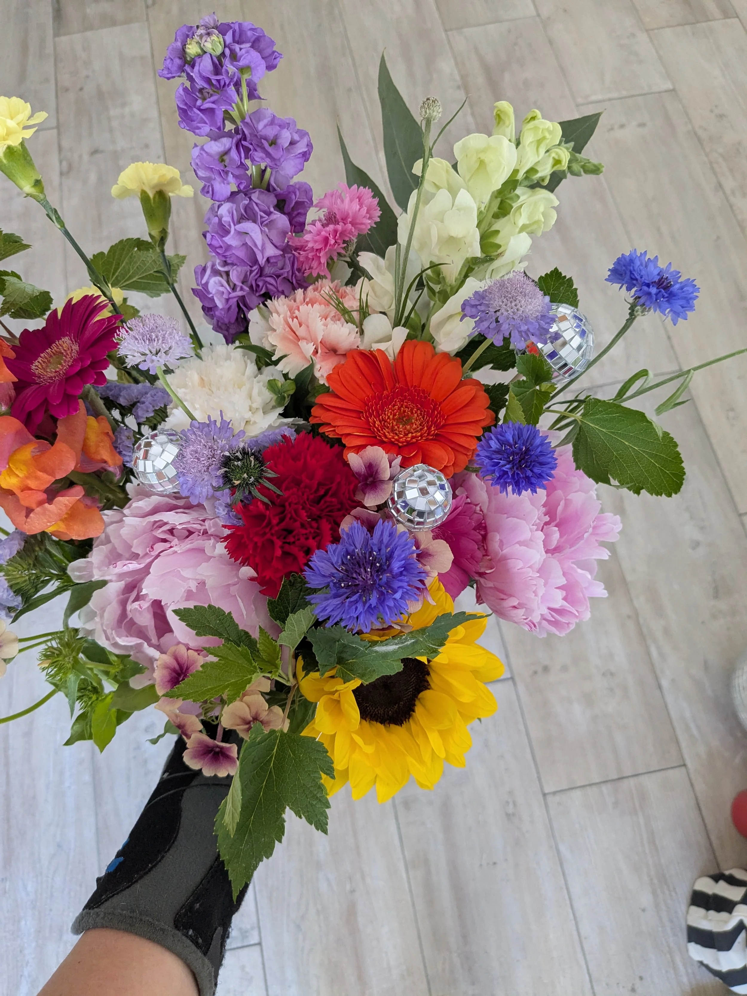 Un bouquet de fleurs variées et colorées, comprenant des lys violets, des gerberas orange, des soucis jaunes, des coquelicots rouges, et d'autres fleurs, avec des feuilles vertes et quelques décorations en miroir.