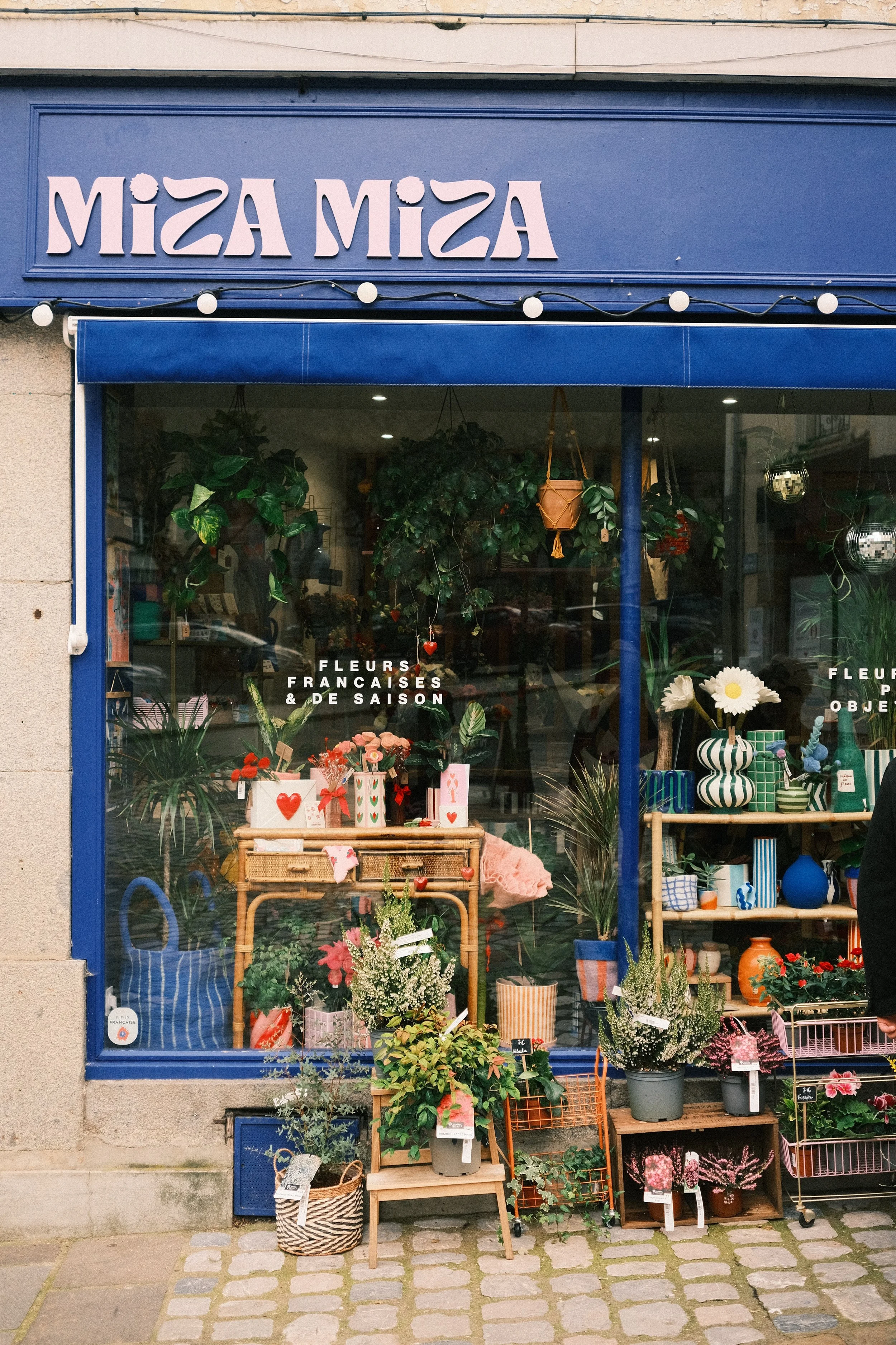 Vitrine d'une boutique de fleurs avec des plantes, bouquets et objets décoratifs. Signage en haut avec le nom 'MIZA MIZA'.