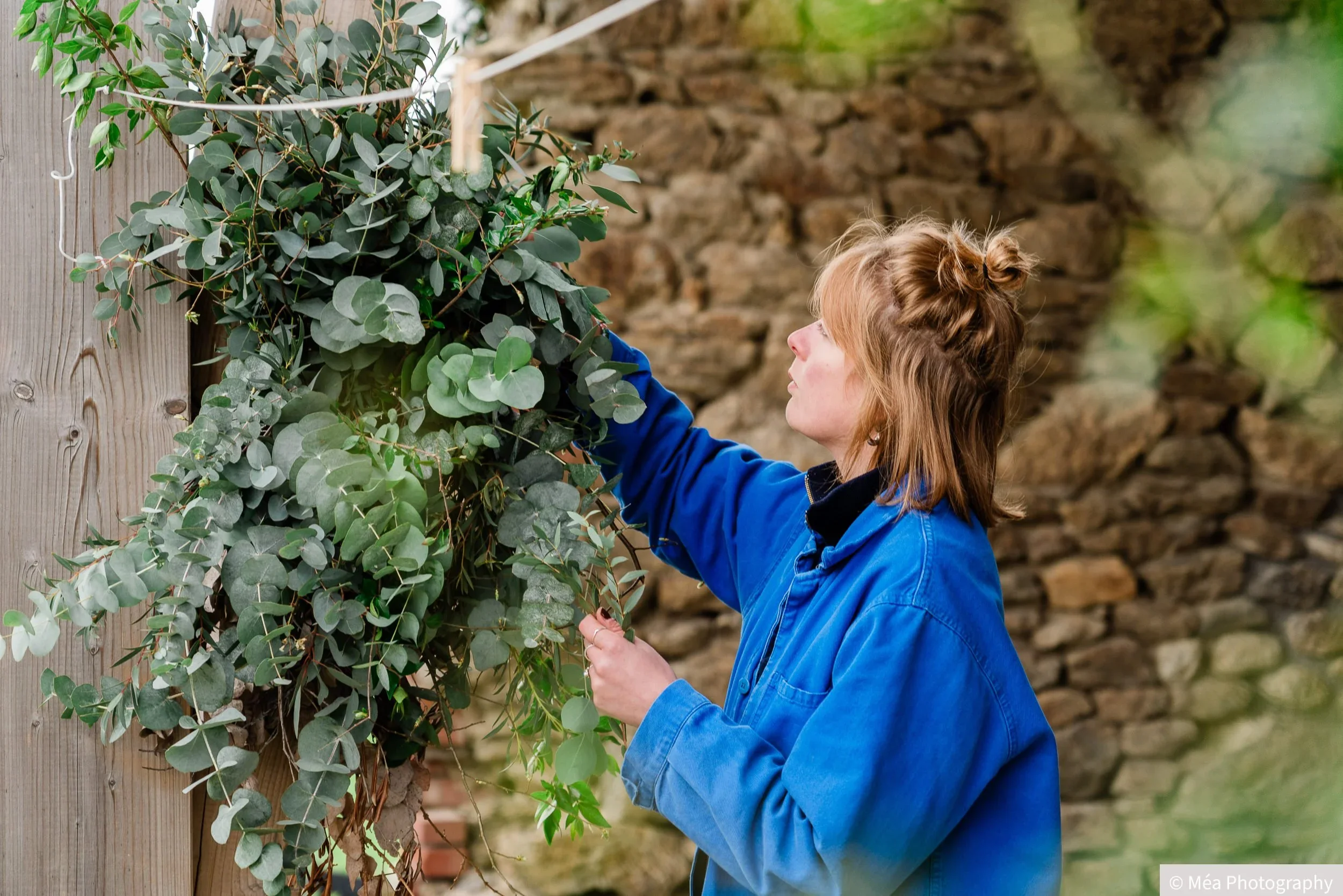 Leslie la fleuriste, portant une veste bleue, coupe des branches ou des feuilles d'une plante verte contre un mur en pierre.