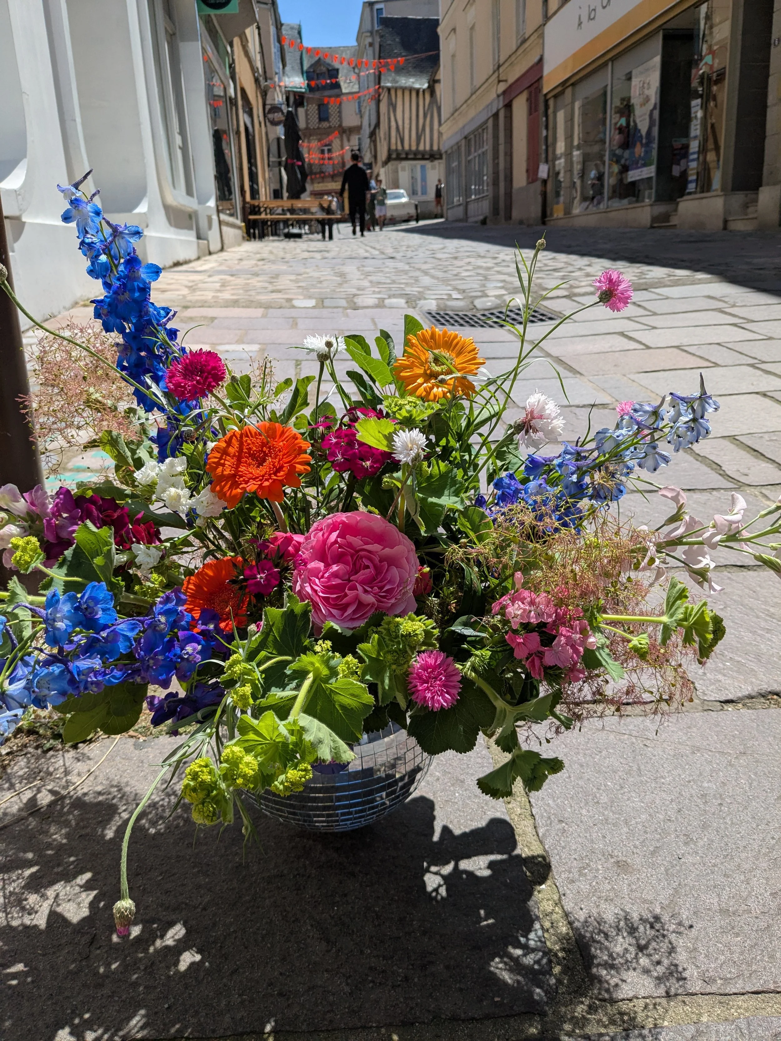 Fleurs colorées dans un panier en osier sur le trottoir d'une rue pavée ensoleillée, avec des bâtiments en arrière-plan et des personnes qui marchent.