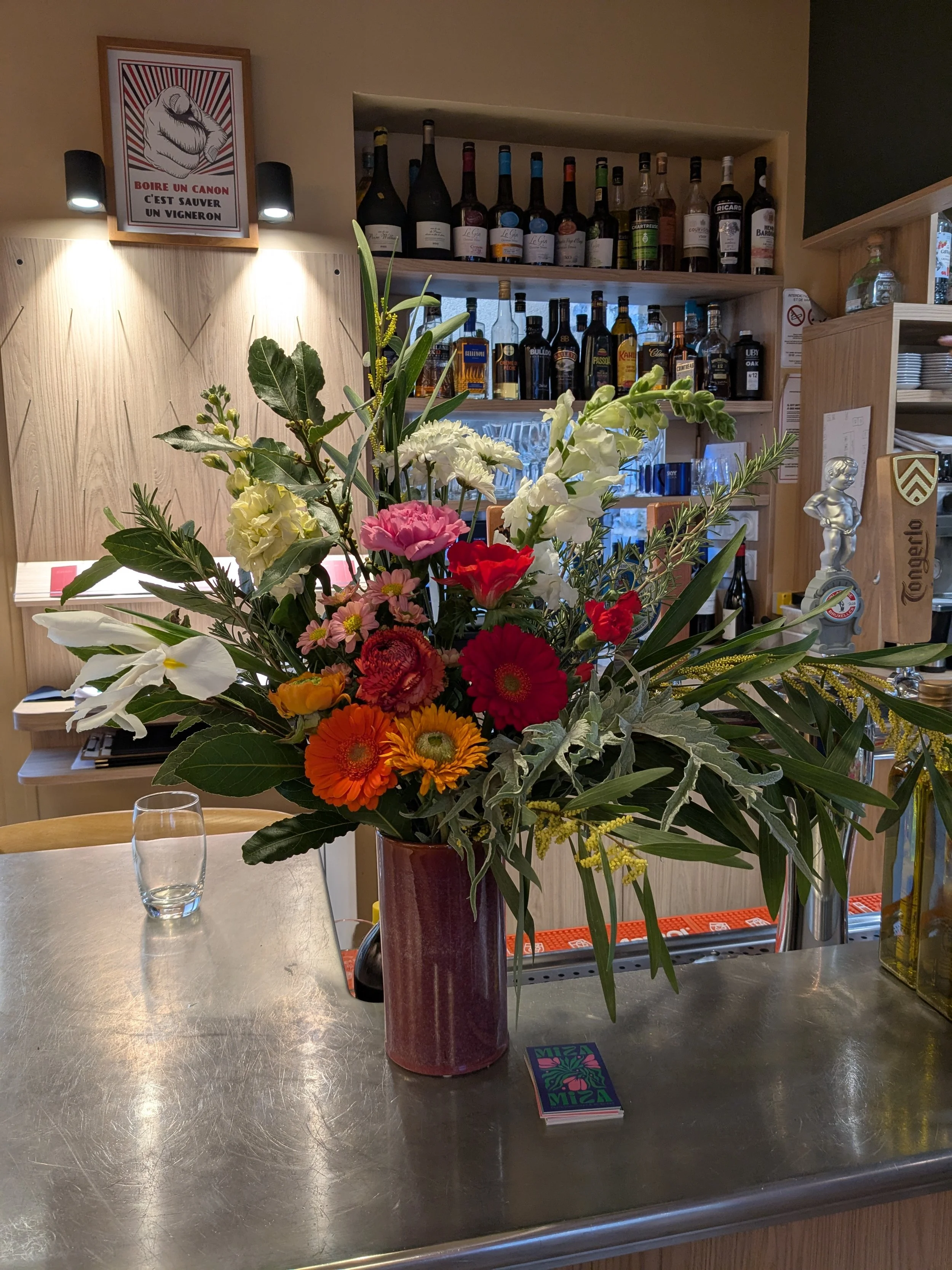 Un grand bouquet de fleurs colorées en vase dans un restaurant ou bar, avec un fond de étagères de bouteilles de vin et d'alcool, et un tableau avec une main pointant du doigt.
