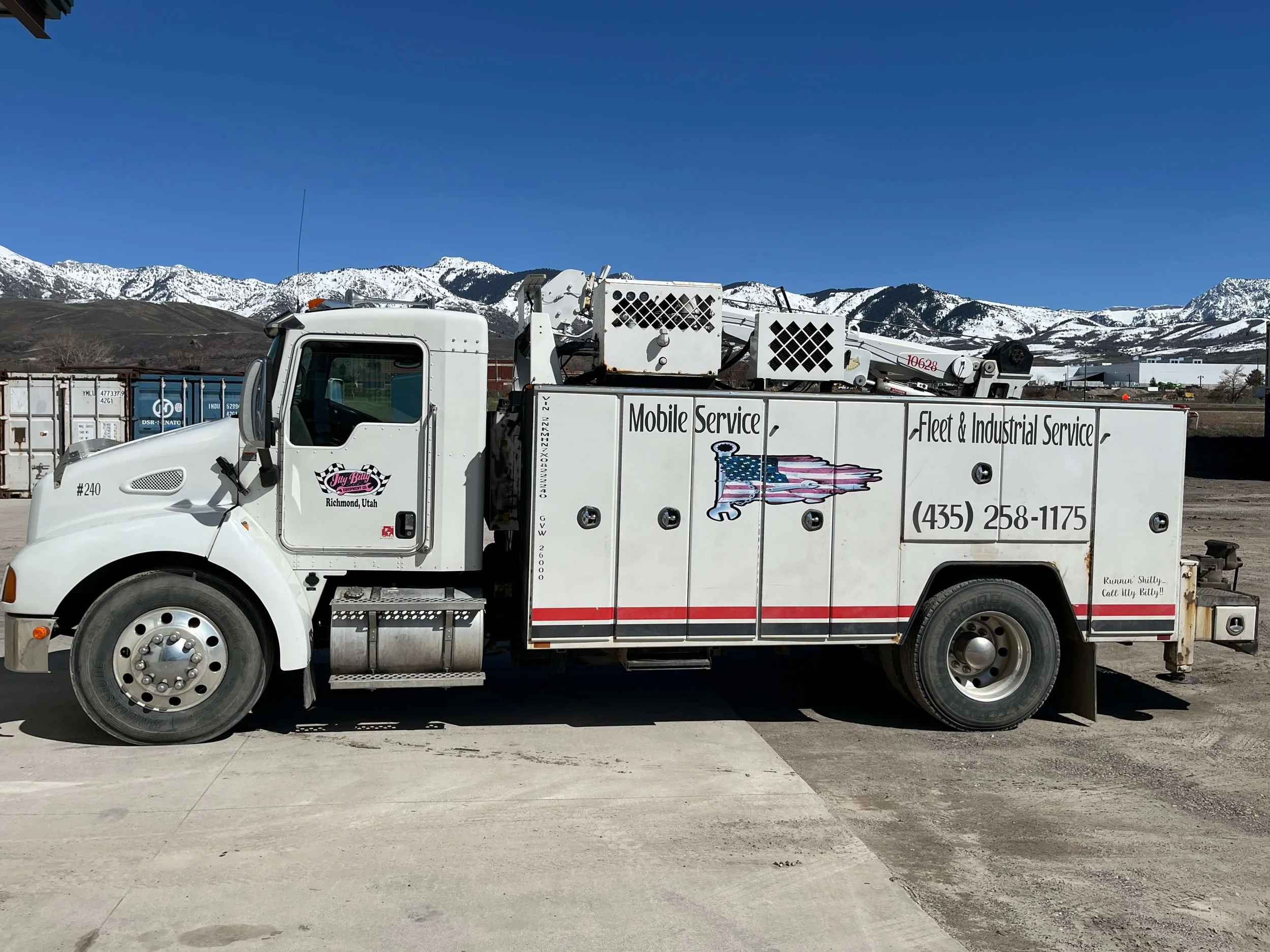 A white mobile service truck with signage for fleet and industrial services, parked on a concrete surface with snow-covered mountains in the background.