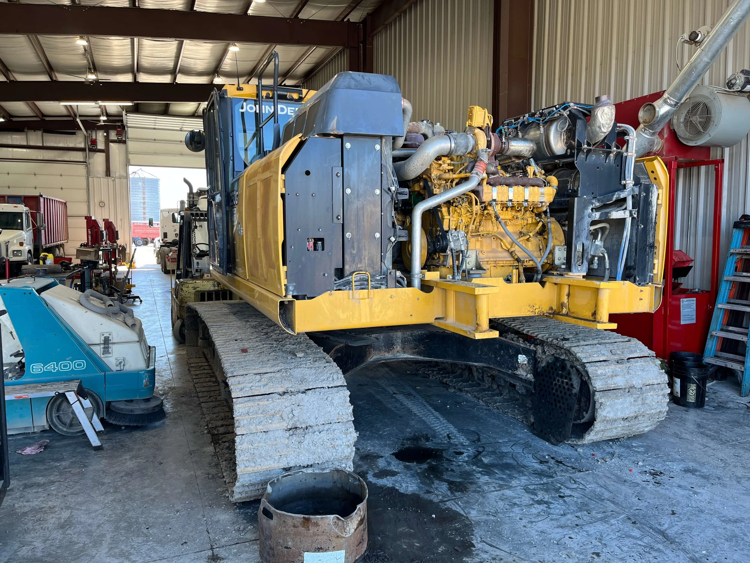 Large yellow construction vehicle, likely an excavator, with exposed engine, inside a garage with industrial equipment and vehicles.