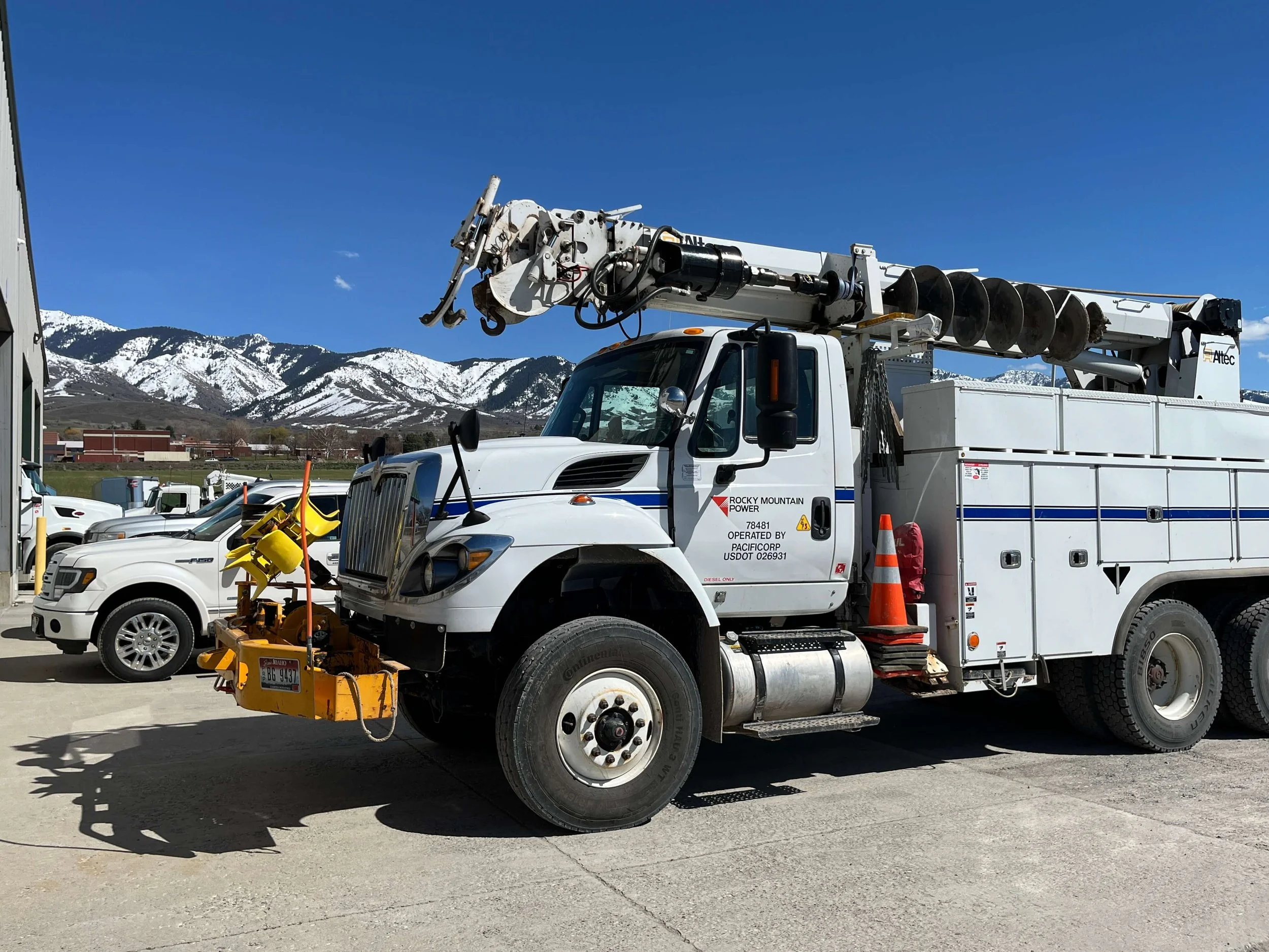 Utility truck with boom and auger parked near other vehicles, with snow-covered mountains in the background.