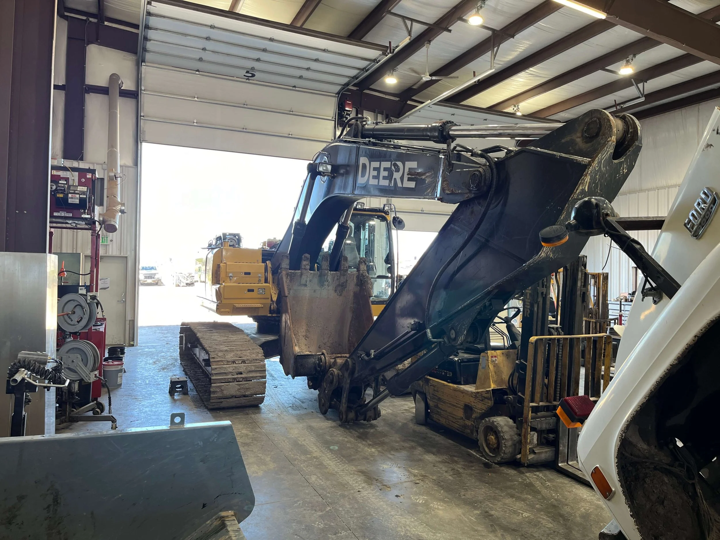 Interior of a garage with a large excavator and forklift, Ford vehicle, and tools, under a metal roof.