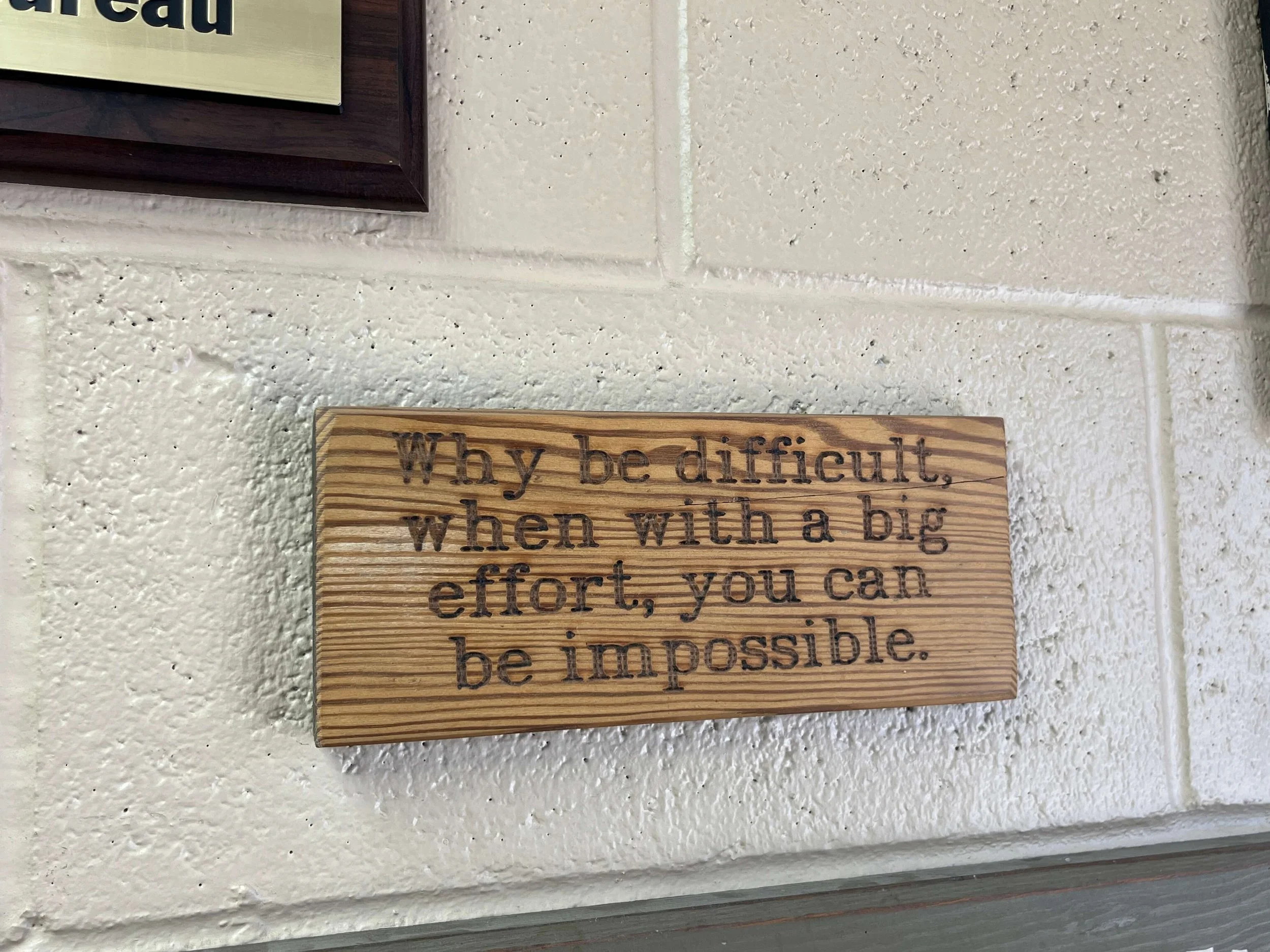 Wooden plaque with the text "Why be difficult, when with a big effort, you can be impossible." mounted on a white brick wall.