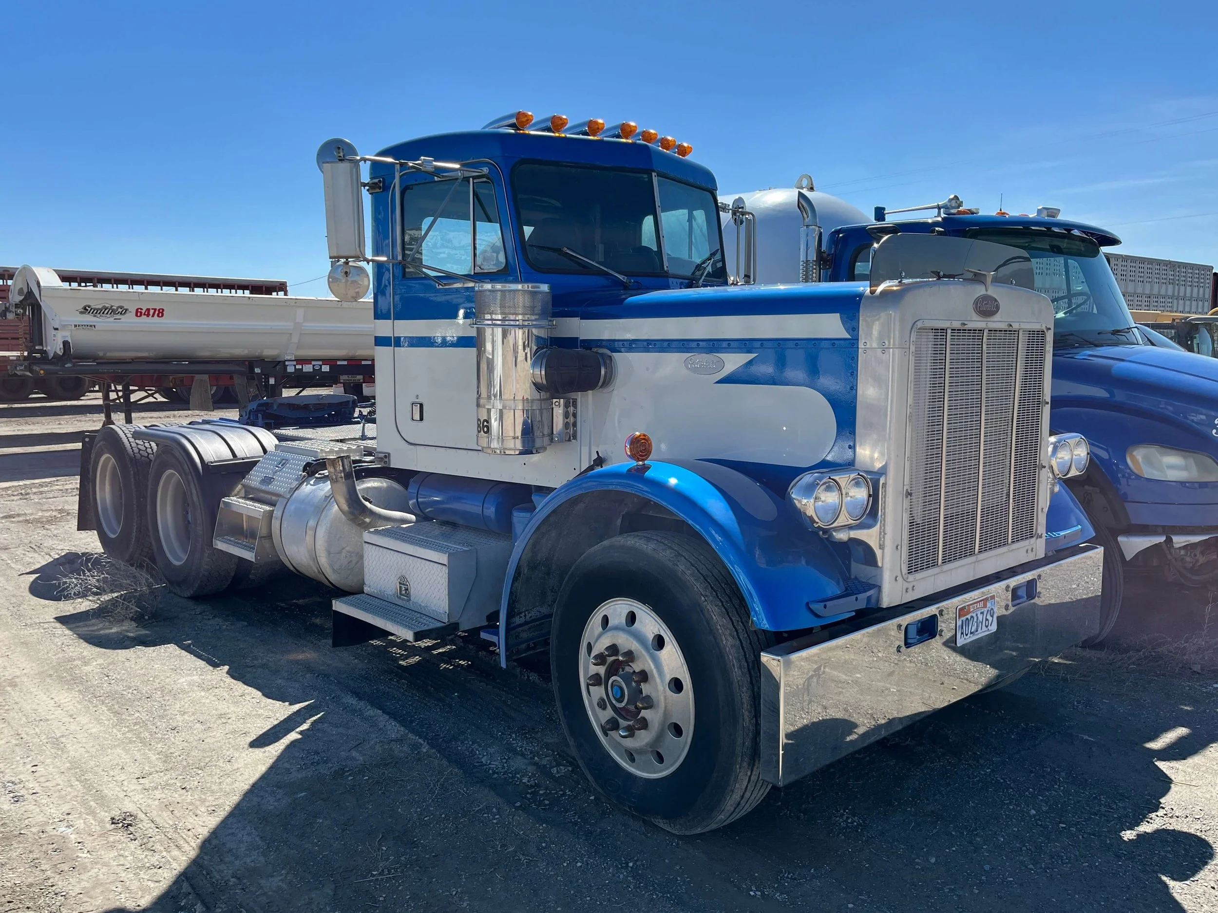 Blue and white semi-truck parked on a gravel lot with a clear blue sky in the background.