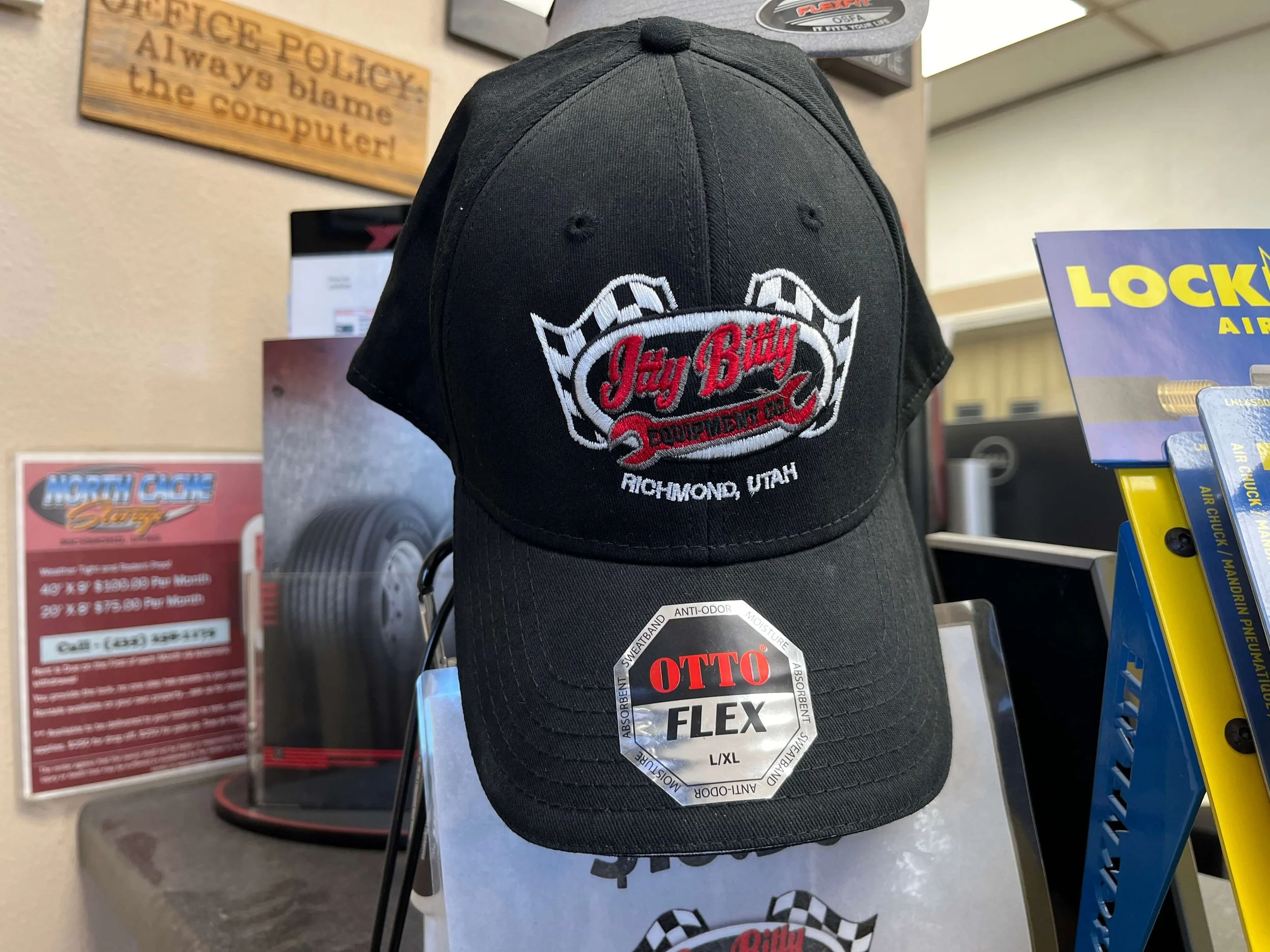 Black baseball cap with "Jay Bitty Equipment Co, Richmond, Utah" logo, displayed on a store shelf next to promotional signs and product boxes.