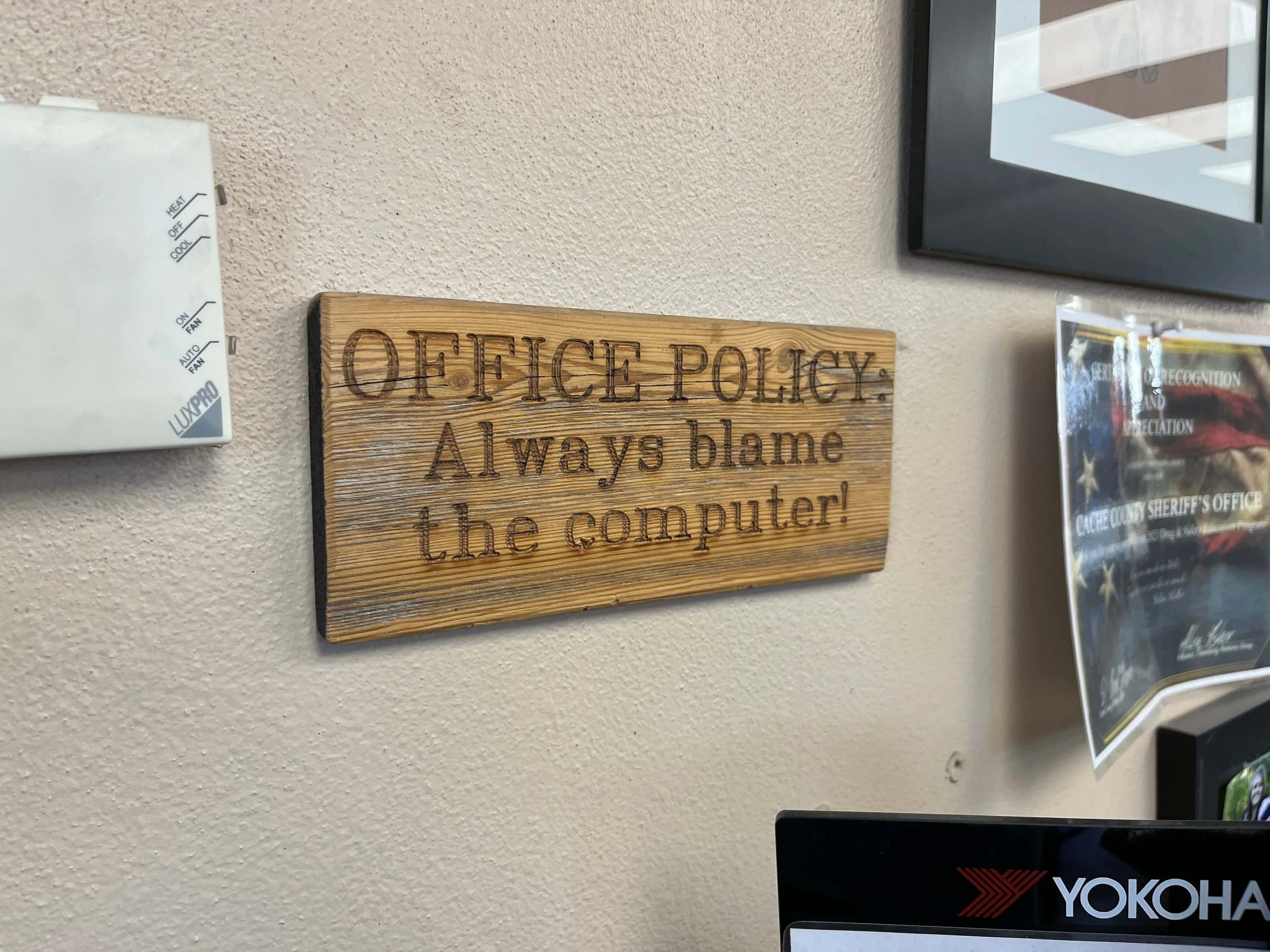 Wooden sign on a wall with the text 'OFFICE POLICY: Always blame the computer!' next to a thermostat and certificates.
