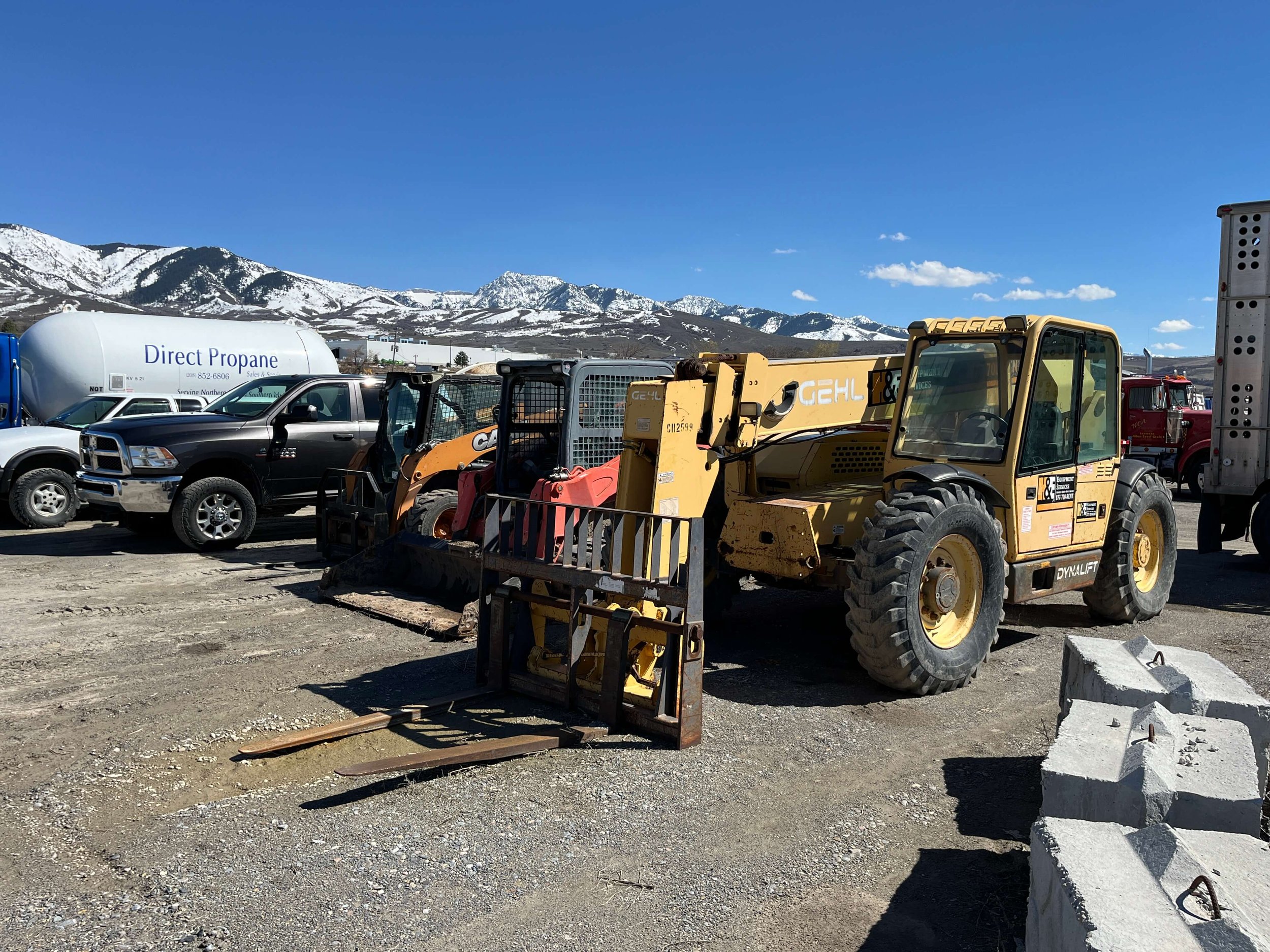 Construction vehicles and trucks parked on a gravel lot with snow-capped mountains in the background.