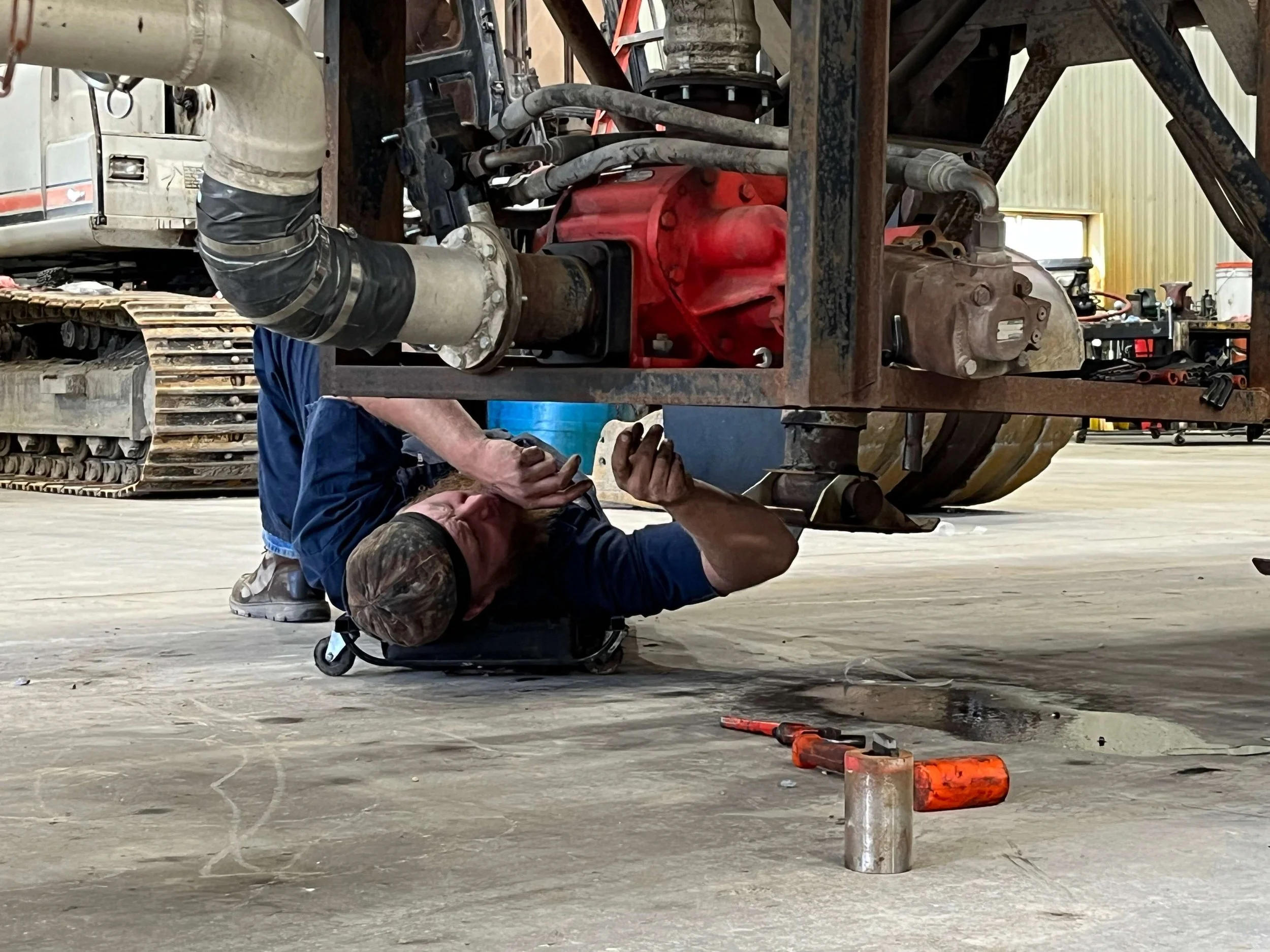 Mechanic working underneath industrial machinery with tools on the floor in a workshop.
